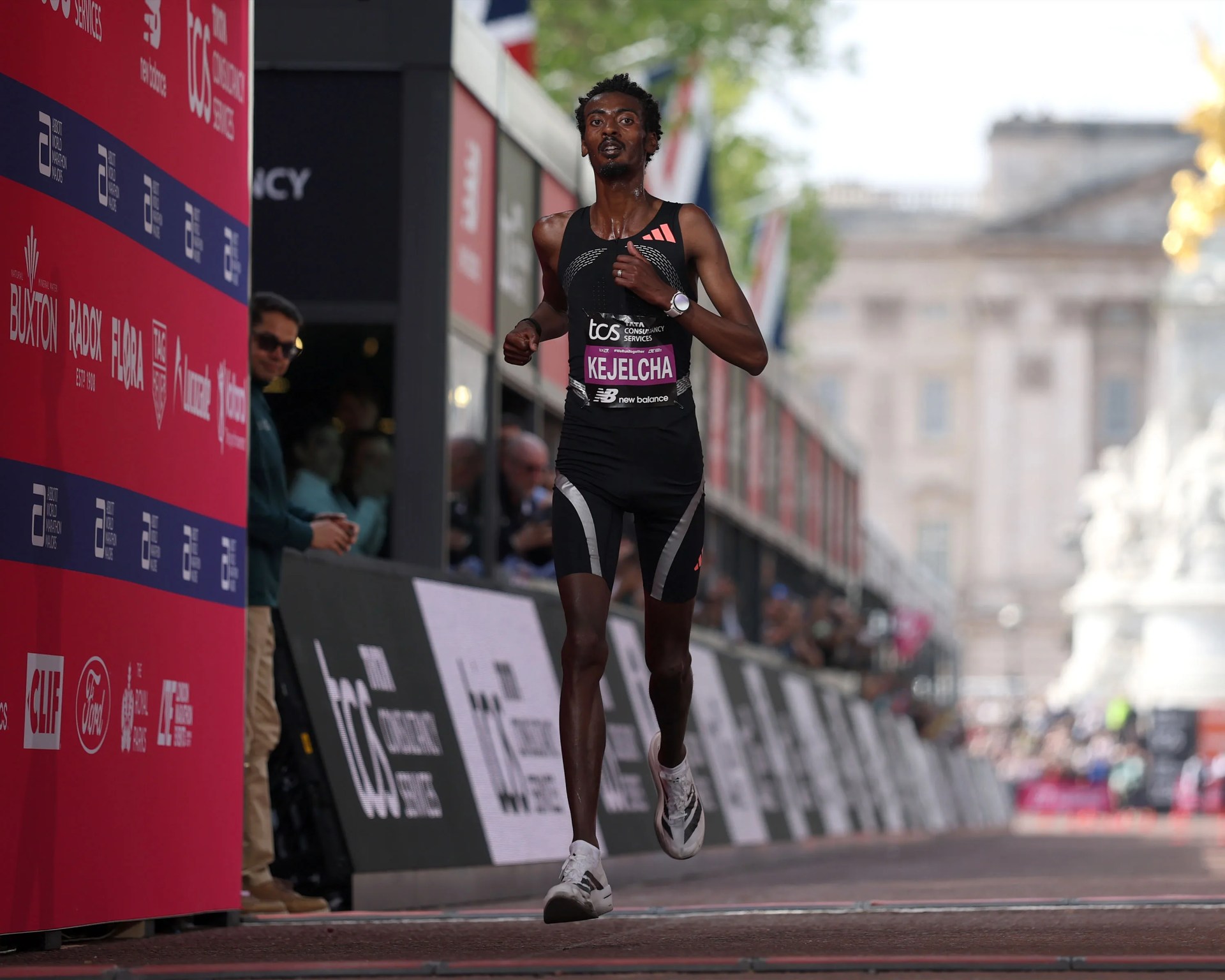 Runner wearing black Adidas racing outfit with "KEJELCHA" bib running on a city street during a marathon event.