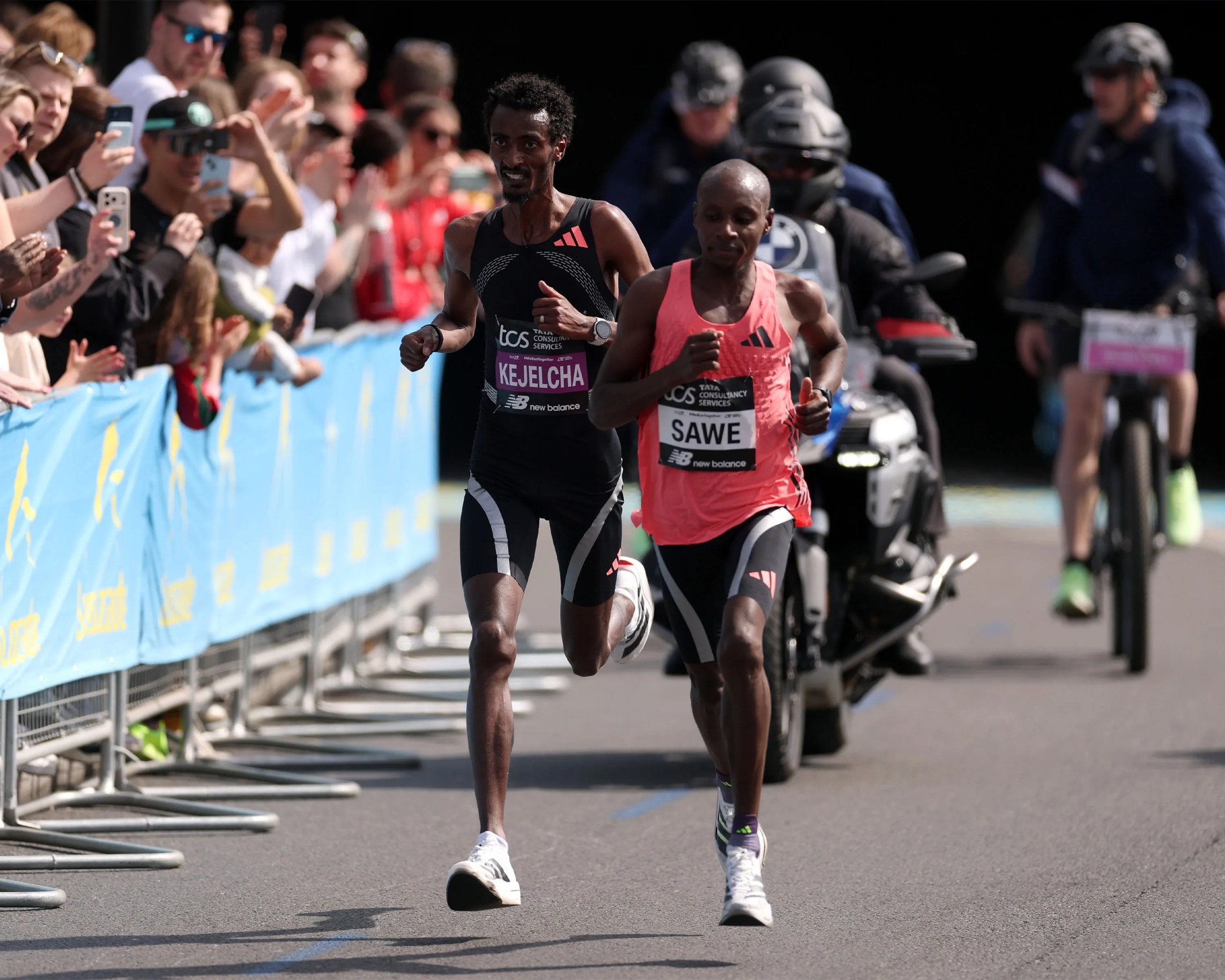 Two male marathon runners in black and pink Adidas racing outfits running on a road with spectators behind blue barriers.