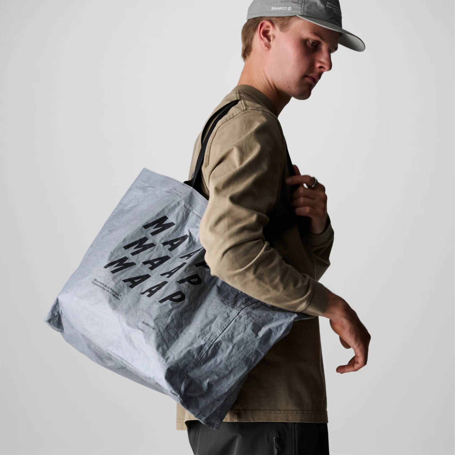 Gray crinkled tote bag with black "MAAP" text carried over shoulder by a person in a brown long-sleeve shirt and gray cap.