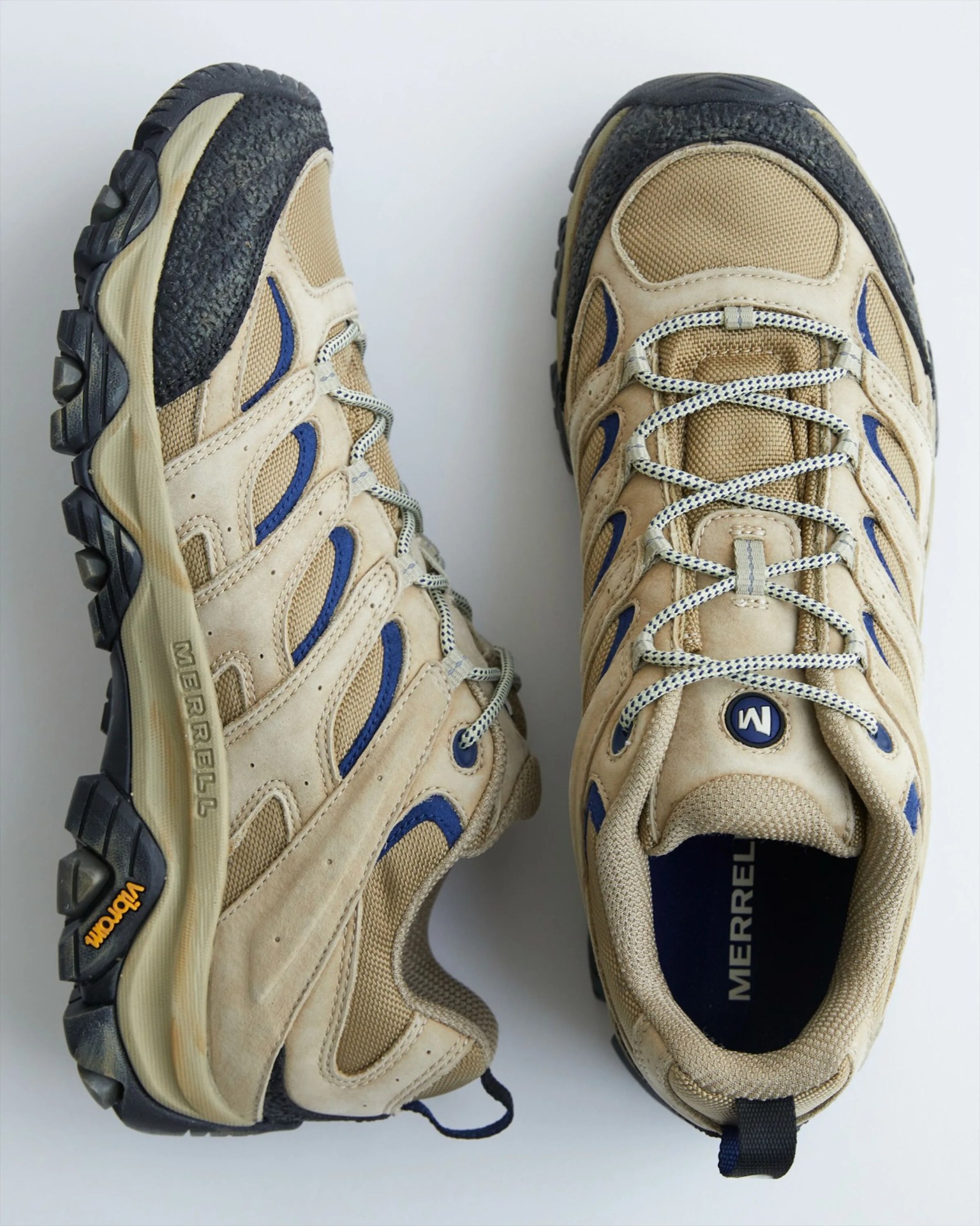 Pair of beige and navy Merrell hiking shoes with black Vibram soles on a white background.