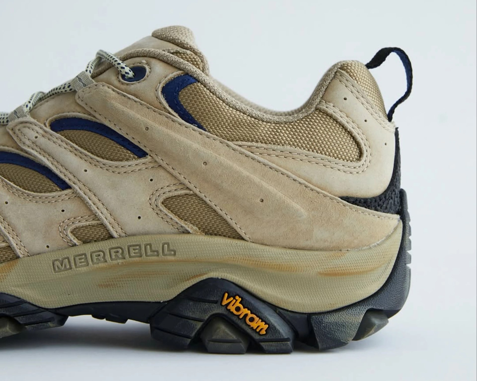 Beige and tan Merrell hiking shoe with black Vibram sole and blue accents on a white background.