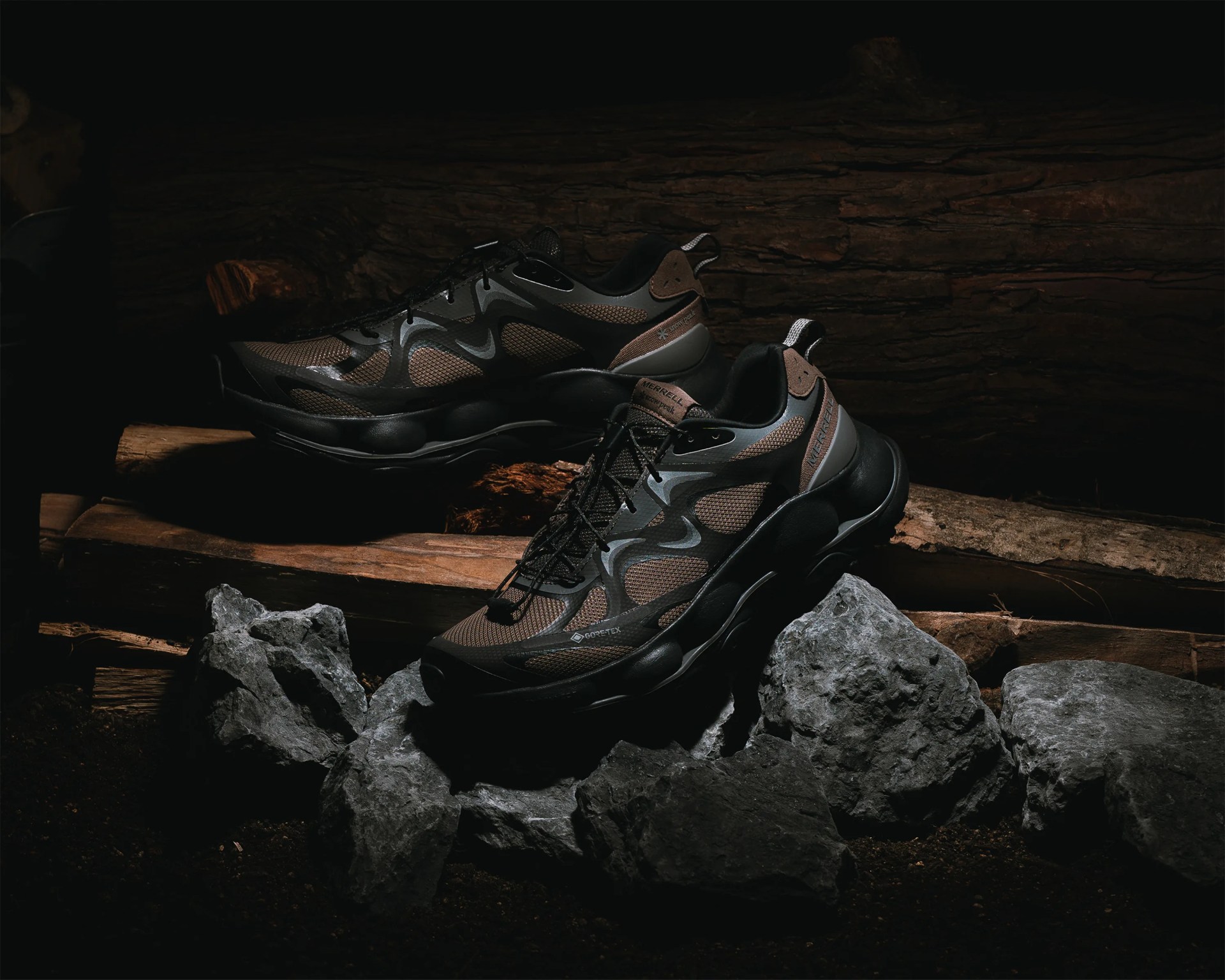 Pair of black and brown Merrell Gore-Tex hiking shoes on gray rocks and wooden logs in low light.