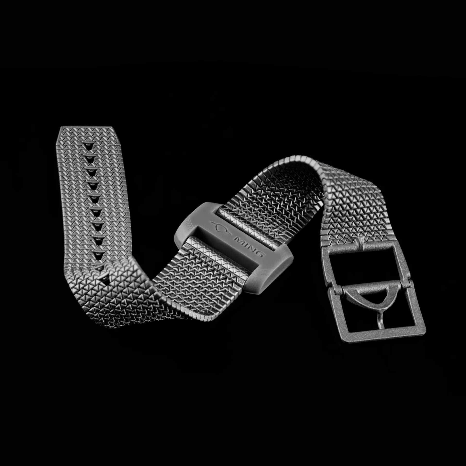 woven metal watch strap with a matte buckle engraved with "MING" on a black background.