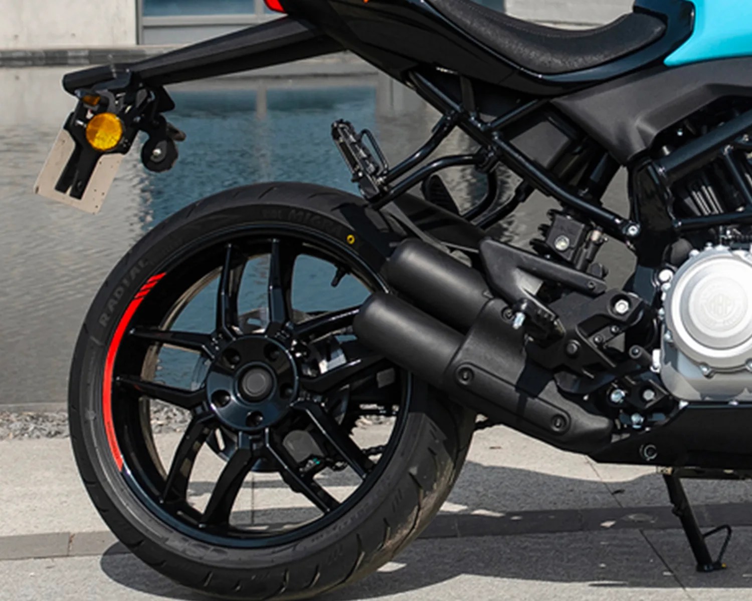 Rear wheel and exhaust pipes of a black motorcycle with red rim accents parked on pavement near water.