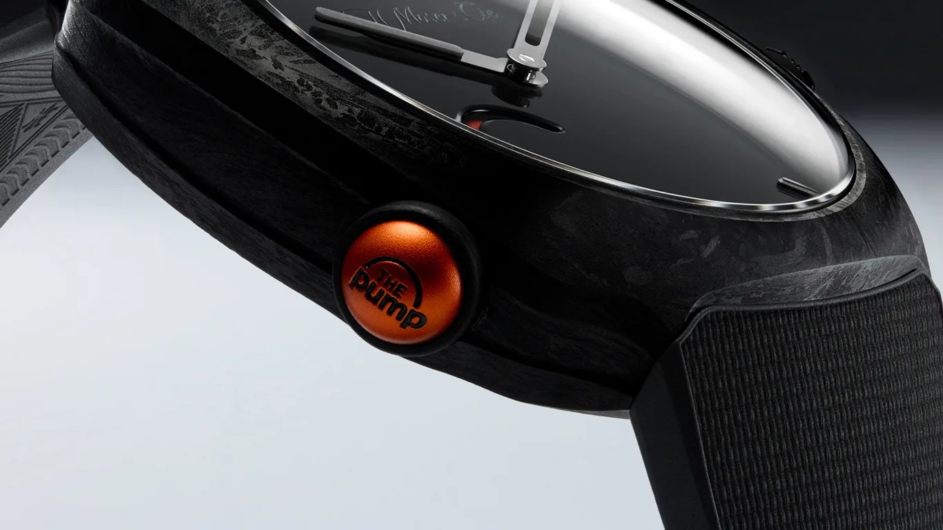 Close-up of a black watch with a textured strap and an orange crown button labeled "THE pump.