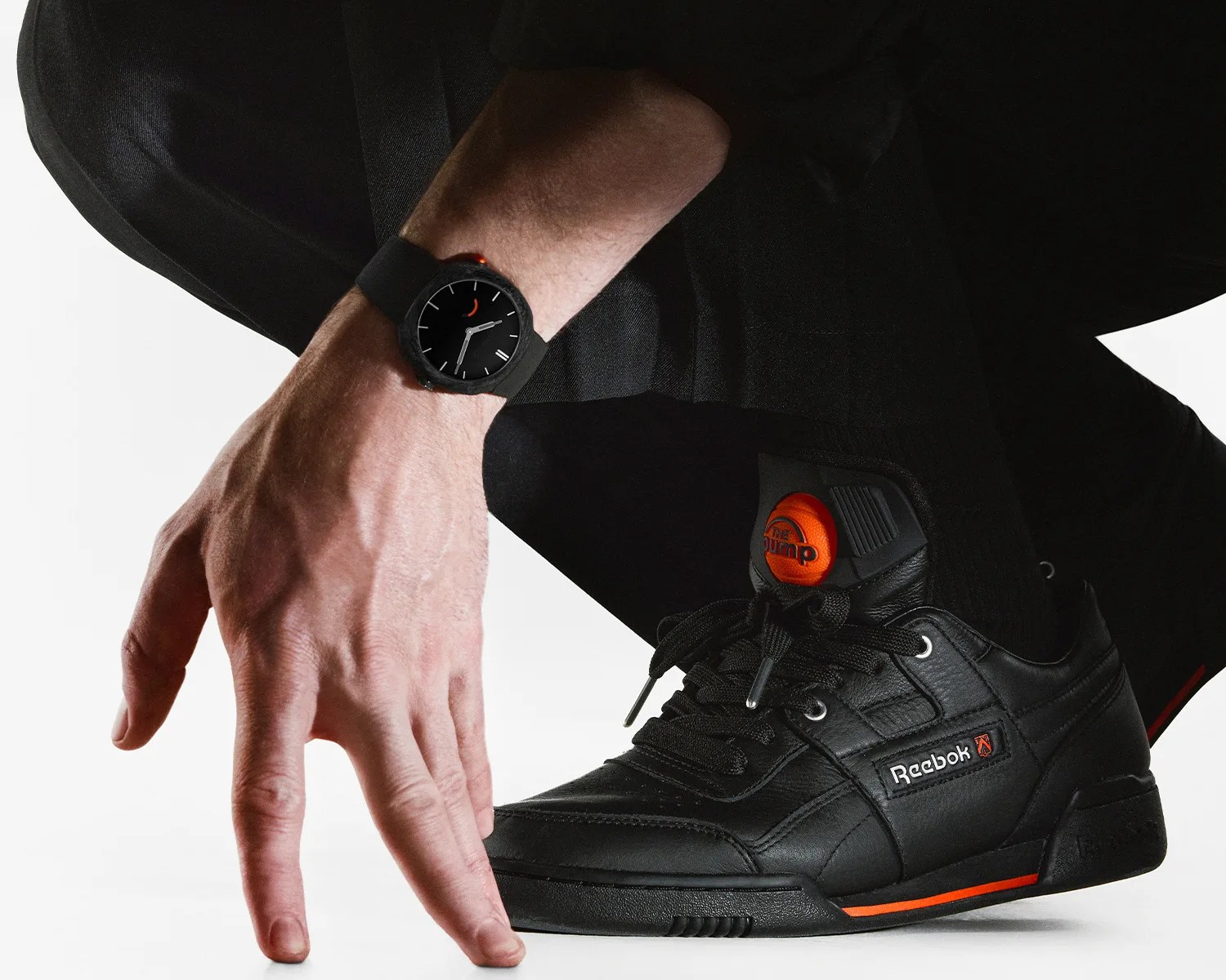 Black Reebok leather sneaker with orange accents and black watch on wrist above it.