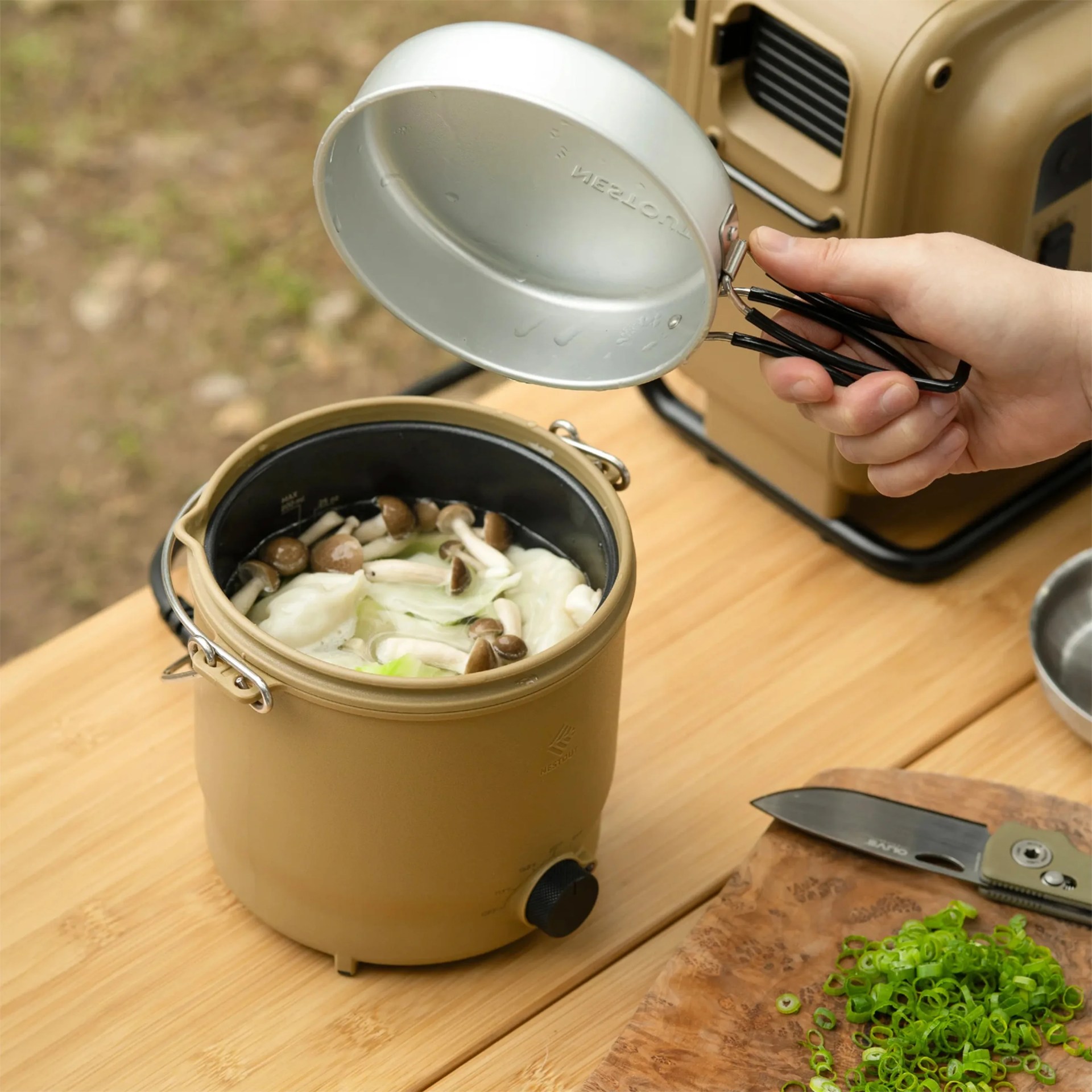 Tan portable electric cooker with a black inner pot containing mushrooms and cabbage, held lid, and chopped green onions on a wooden board with a knife.