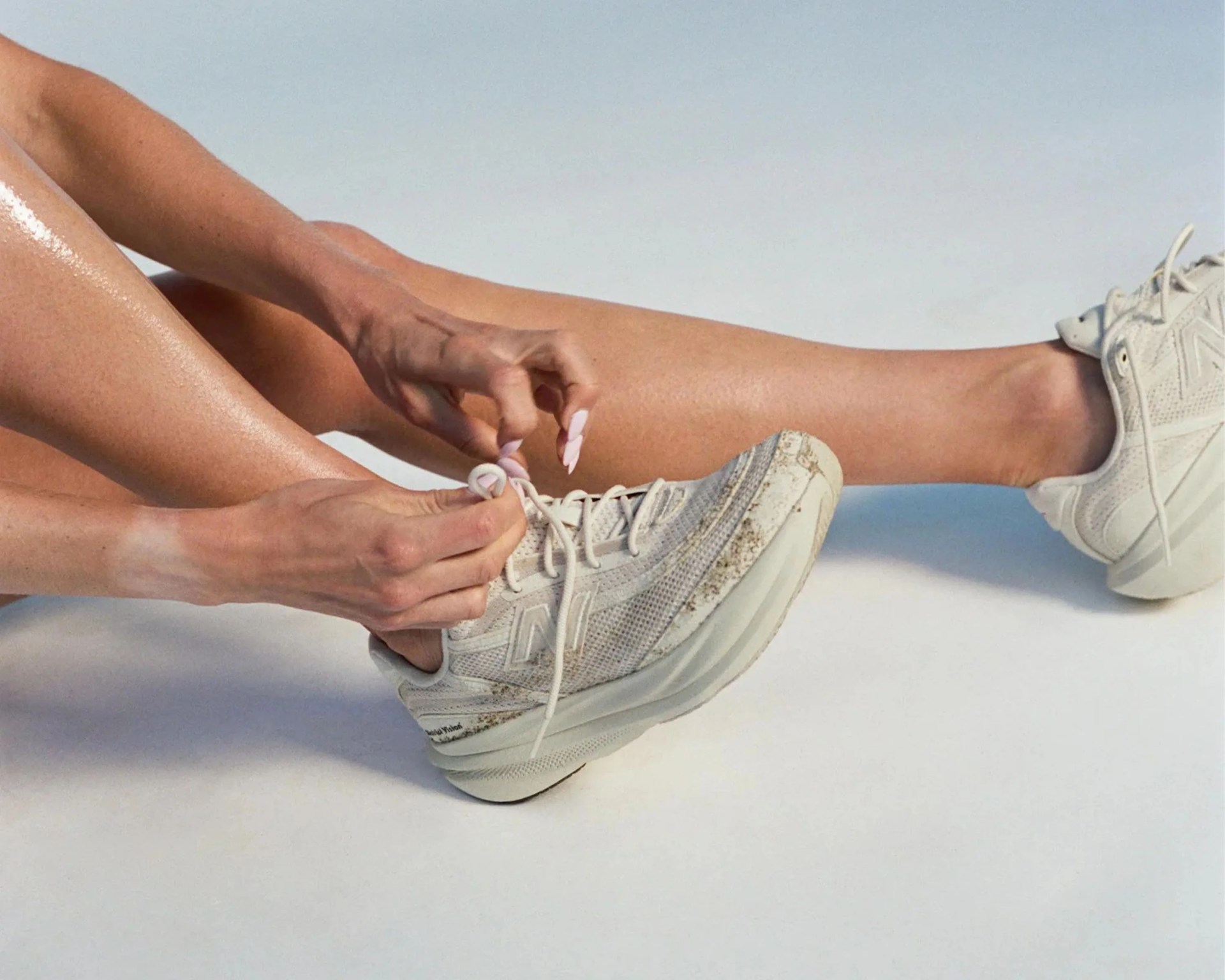 Person tying laces on off-white New Balance sneakers with some dirt marks on a light background.