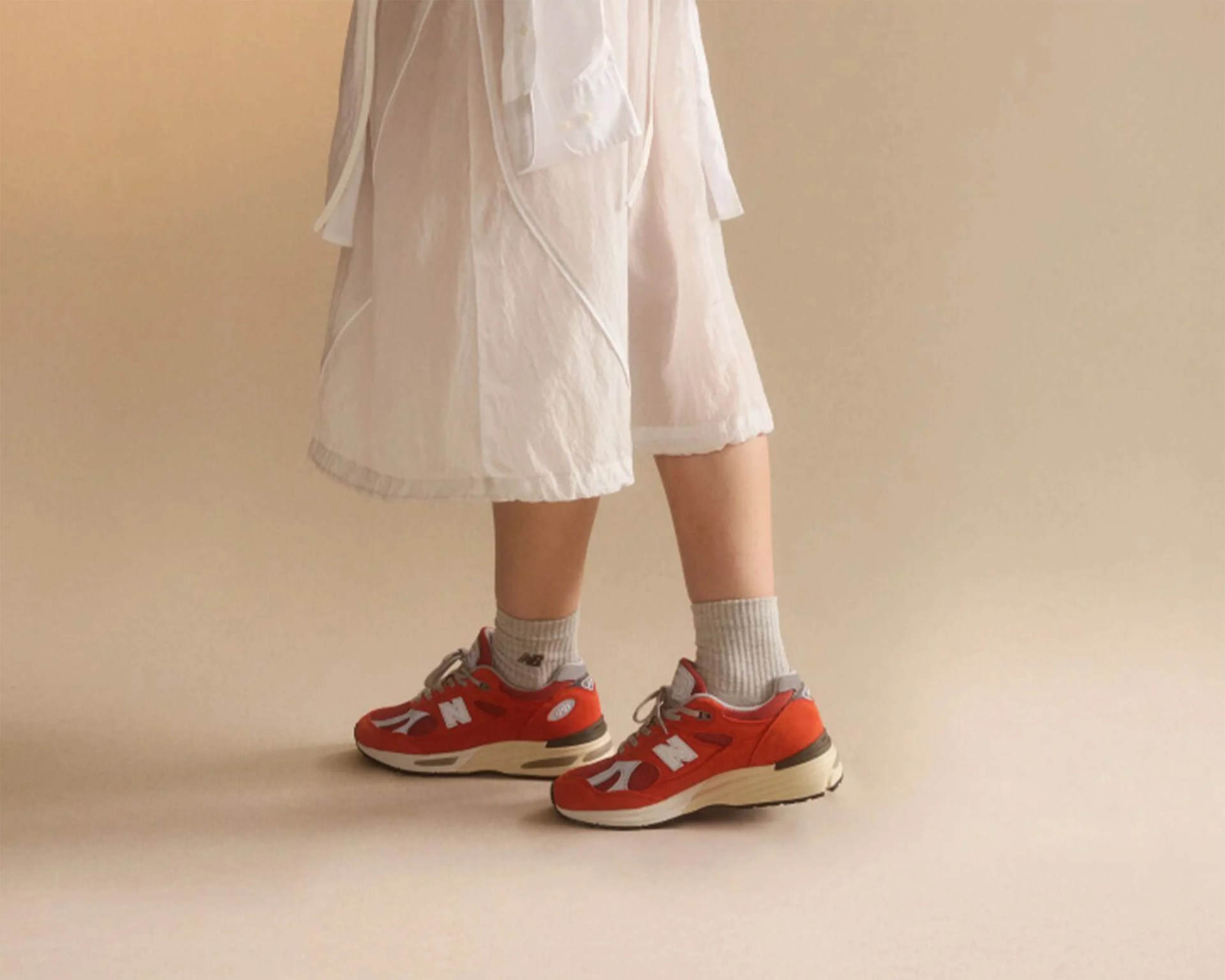 Person wearing beige socks and red New Balance sneakers with white accents and a white skirt.