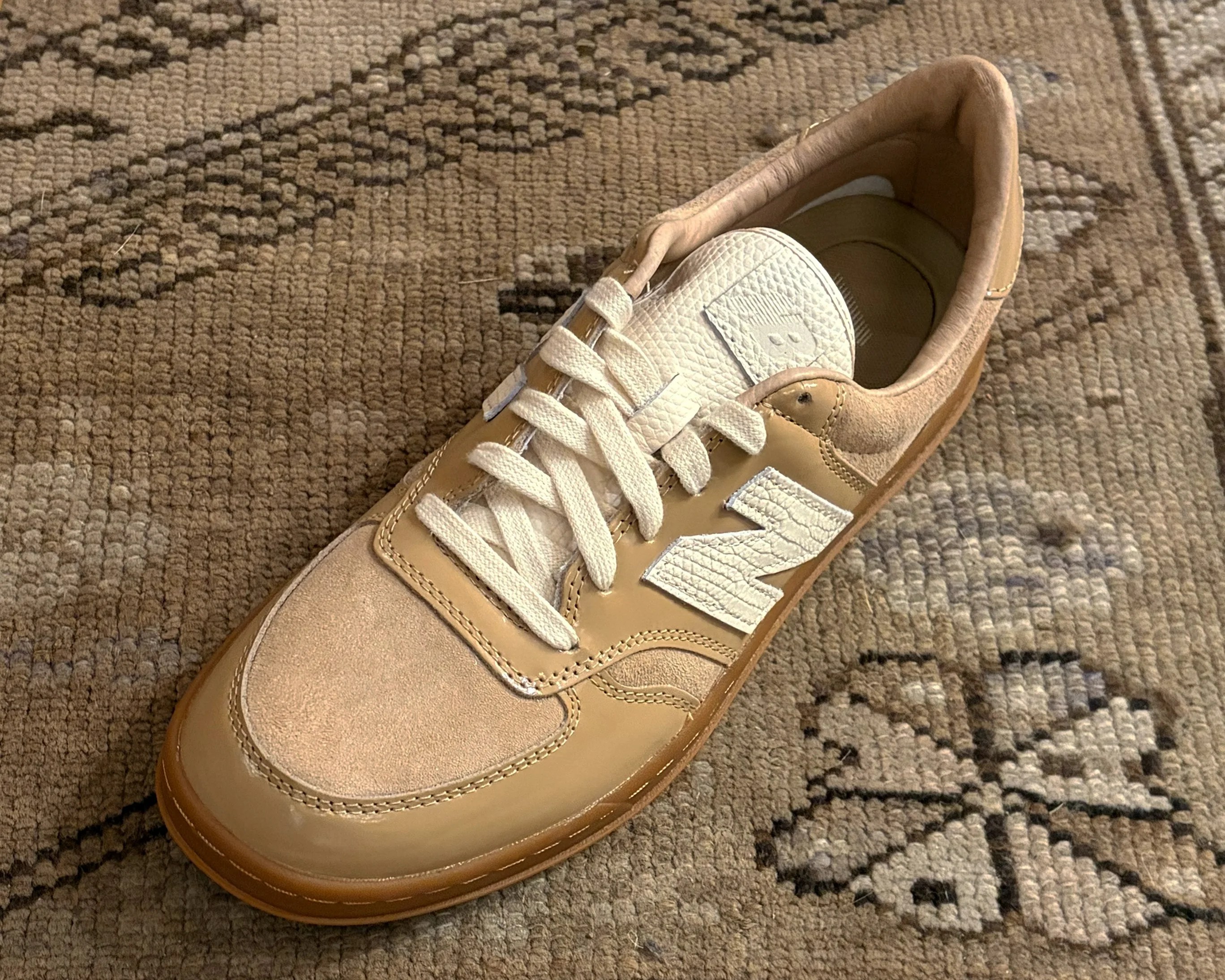 Tan and beige New Balance sneaker with white laces on a patterned brown rug.