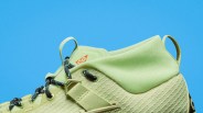 Close-up of a light green sneaker with black and white laces and a small orange logo on the tongue against a blue background.