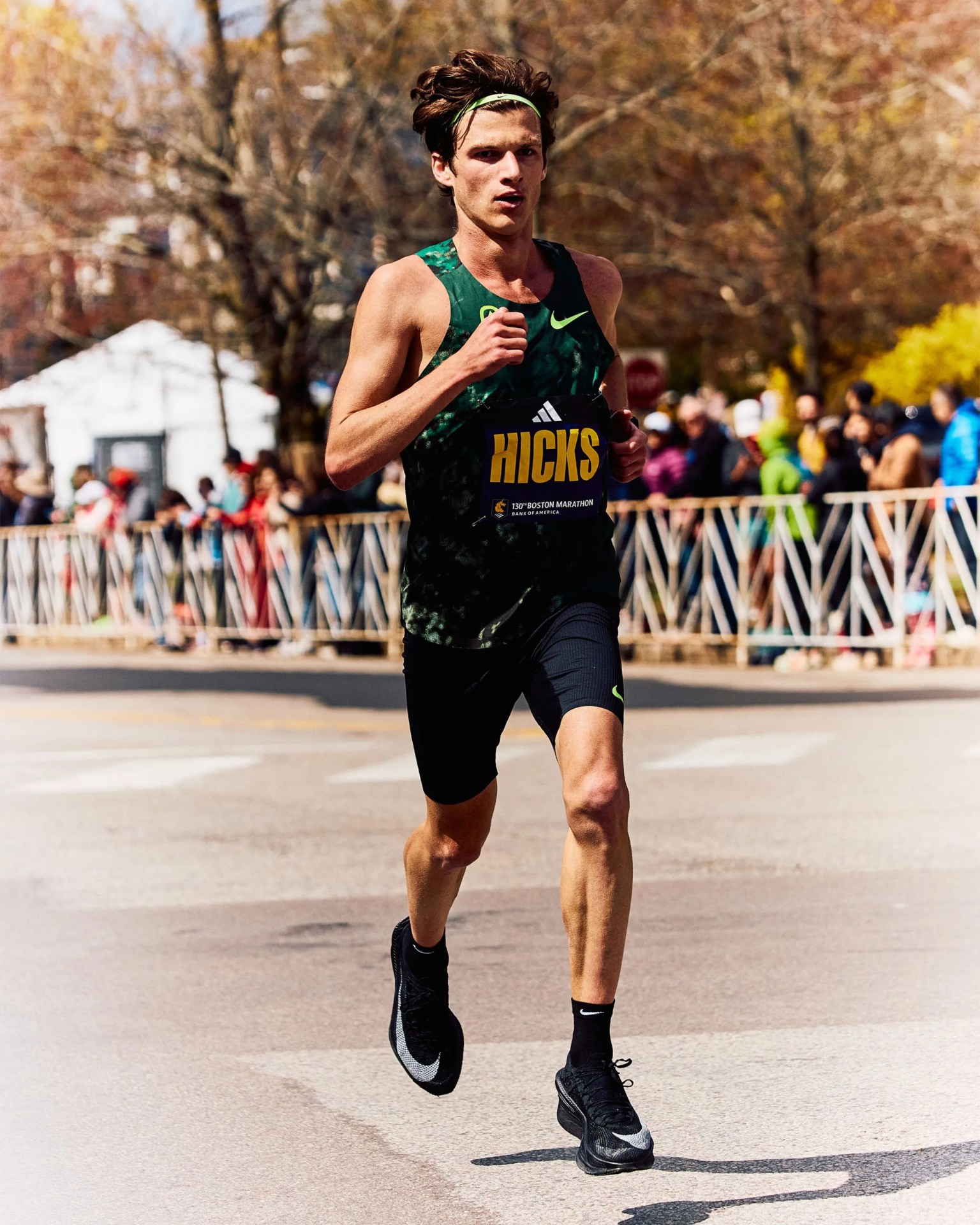 Runner wearing a green and black Nike singlet and black shorts with "HICKS" bib running on a road during a marathon.