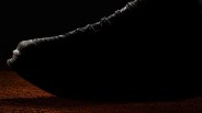 Black textured sneaker with rope-style laces on a dark brown surface against a black background.