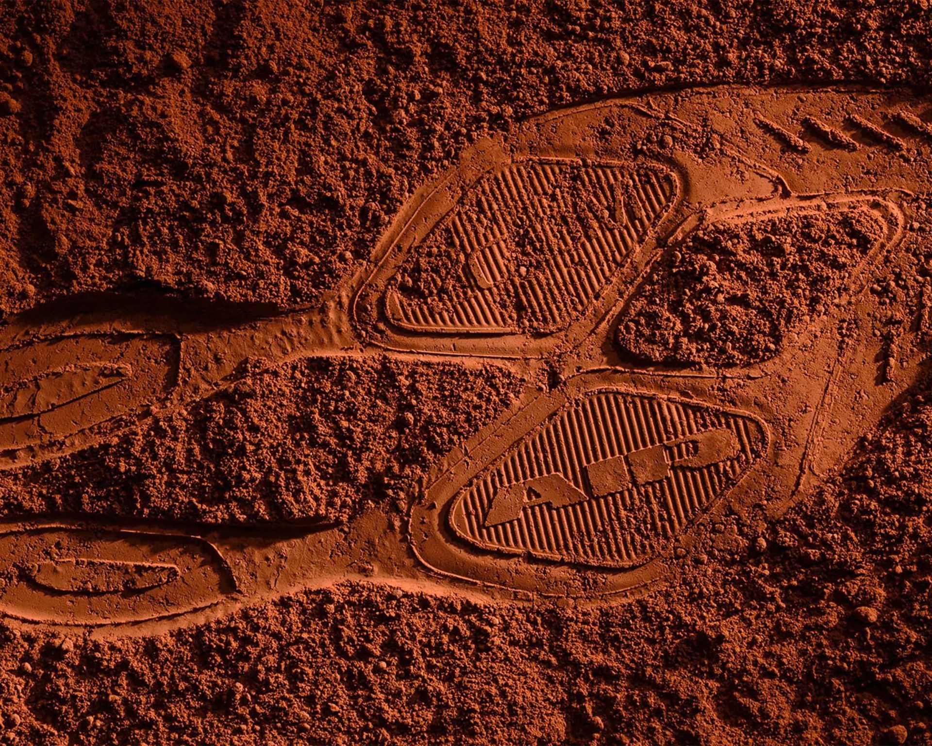 Close-up of a shoe print with detailed tread pattern in reddish-brown soil.