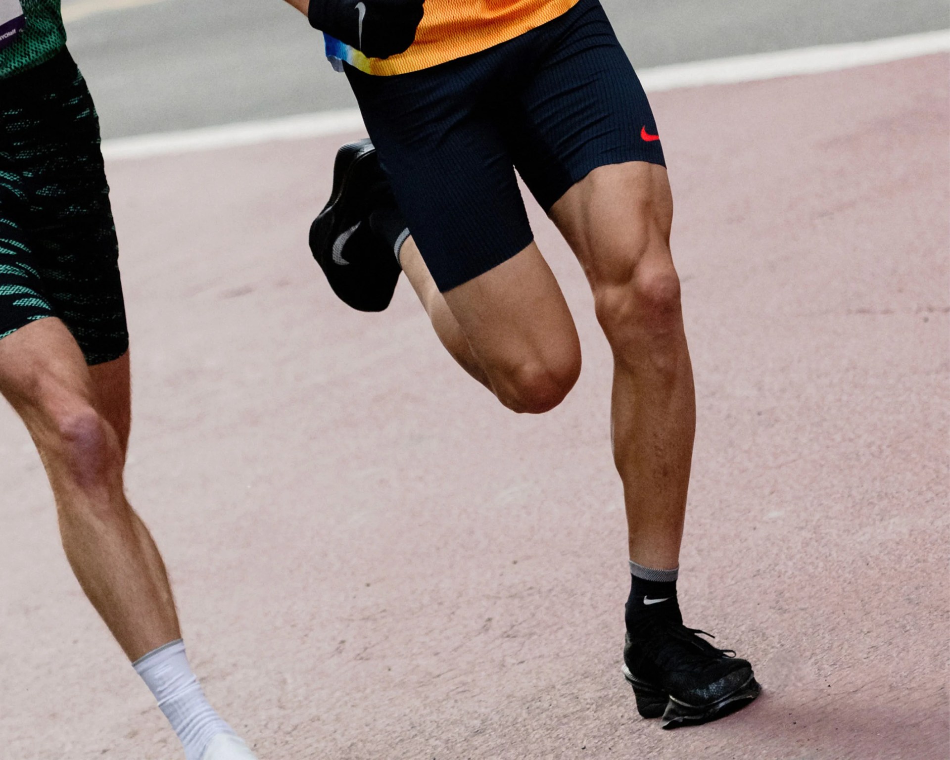 Close-up of two runners' legs in motion on a track, one wearing black Nike shorts and black shoes, the other in green shorts and white shoes.