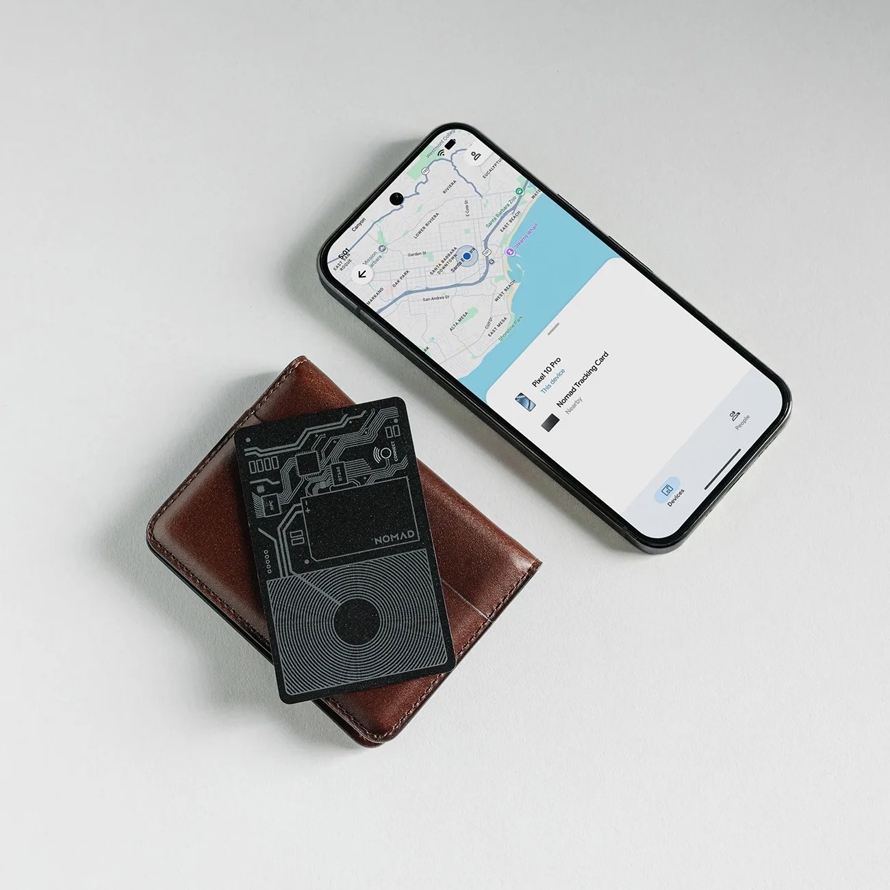 Black Nomad tracking card with circuit design on a brown leather wallet next to a smartphone showing a map.