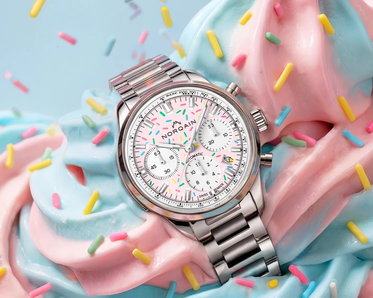 Silver metal wristwatch with a pink dial featuring colorful sprinkles, placed on pastel pink and blue swirled frosting with sprinkles.