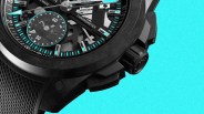 Close-up of a black chronometer watch with turquoise accents and a textured black strap against a bright blue background.