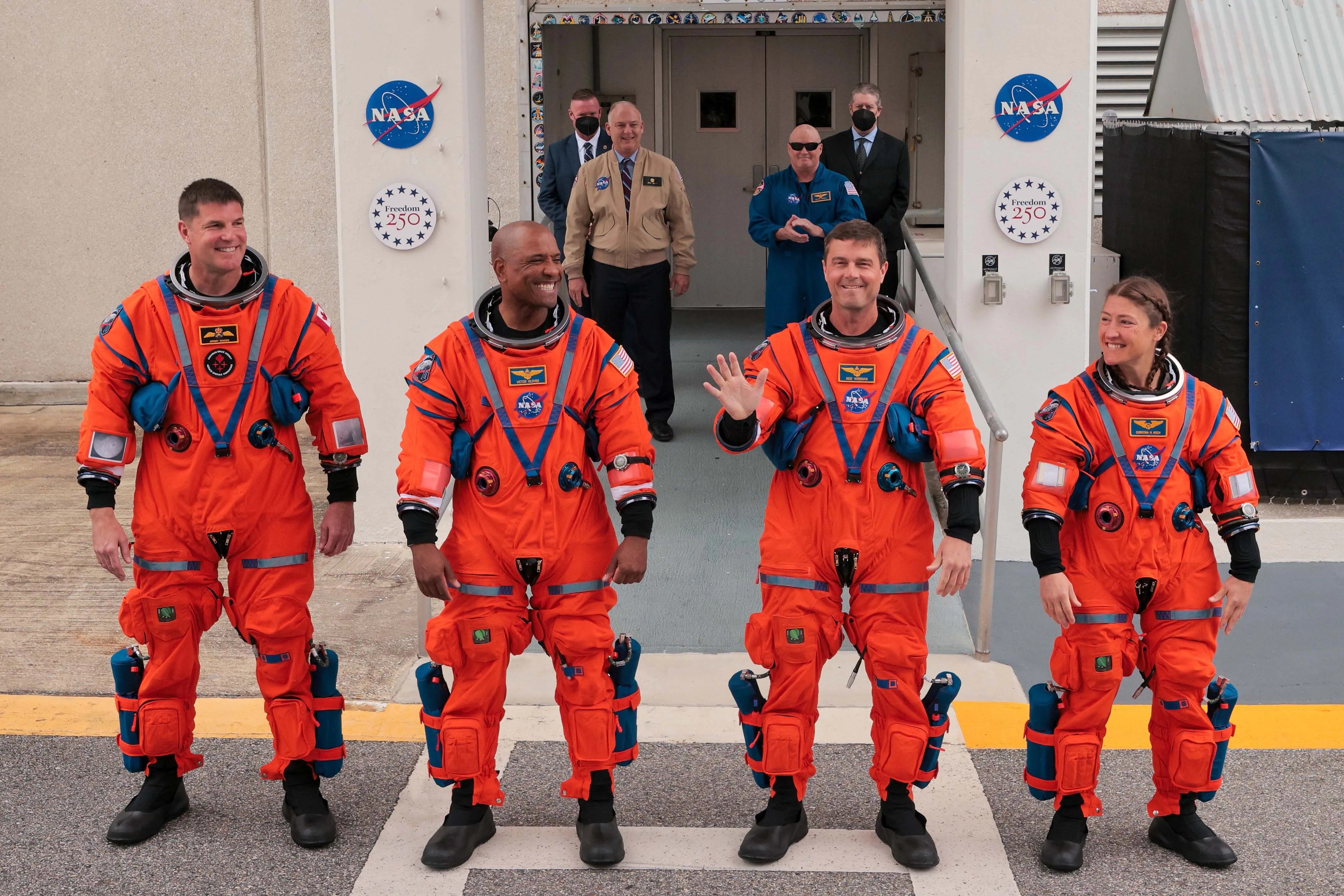 Four astronauts in orange NASA spacesuits standing outside a building with NASA logos and "Freedom 250" signs.