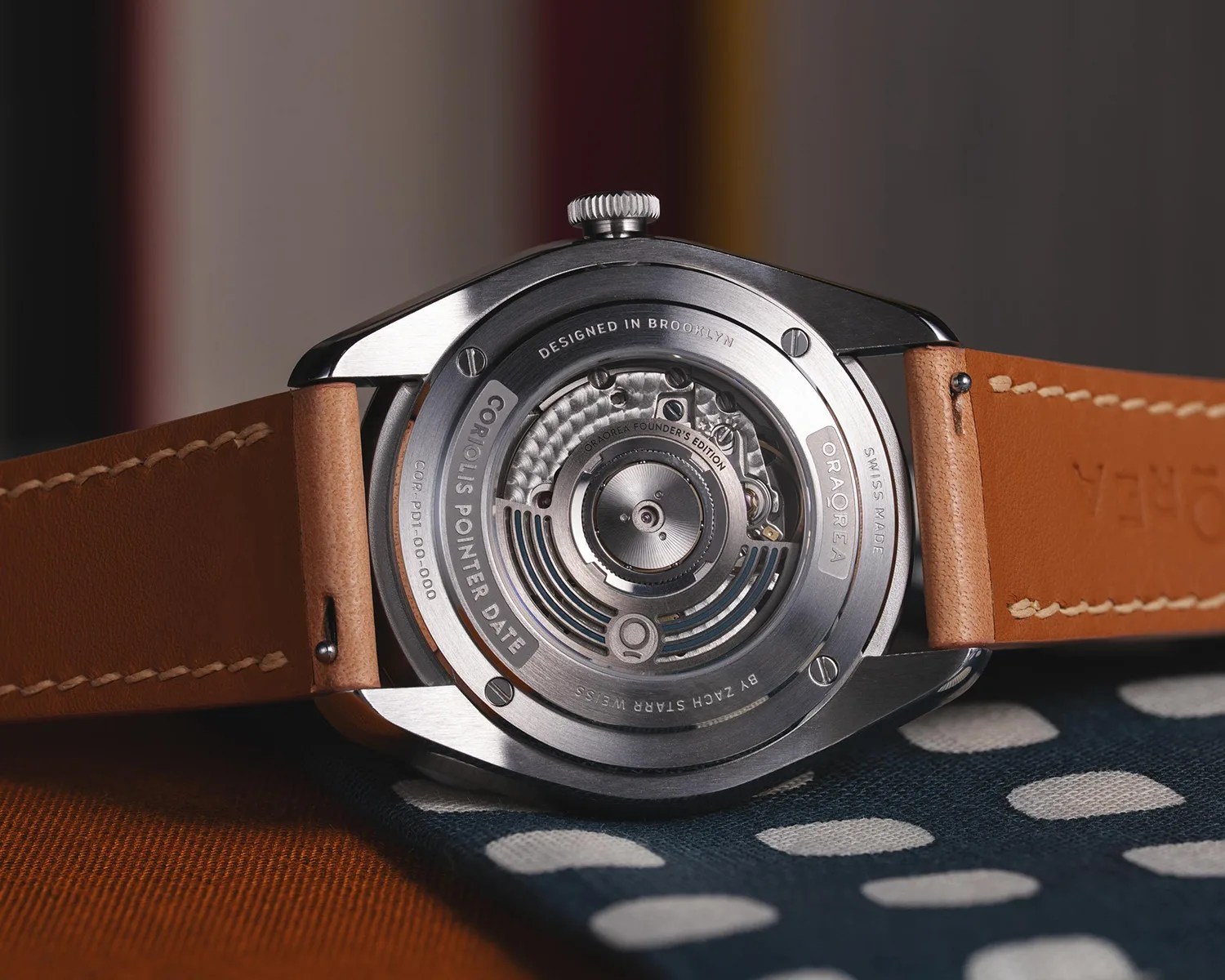 Back of a stainless steel wristwatch with visible mechanical movement and tan leather strap on a patterned fabric.