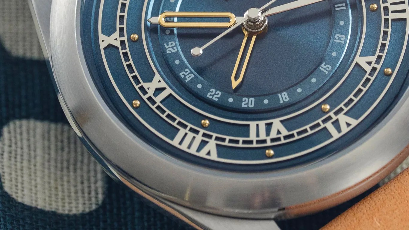 Close-up of a blue watch face with gold and silver hands, Roman numerals, and a silver bezel on a textured surface.