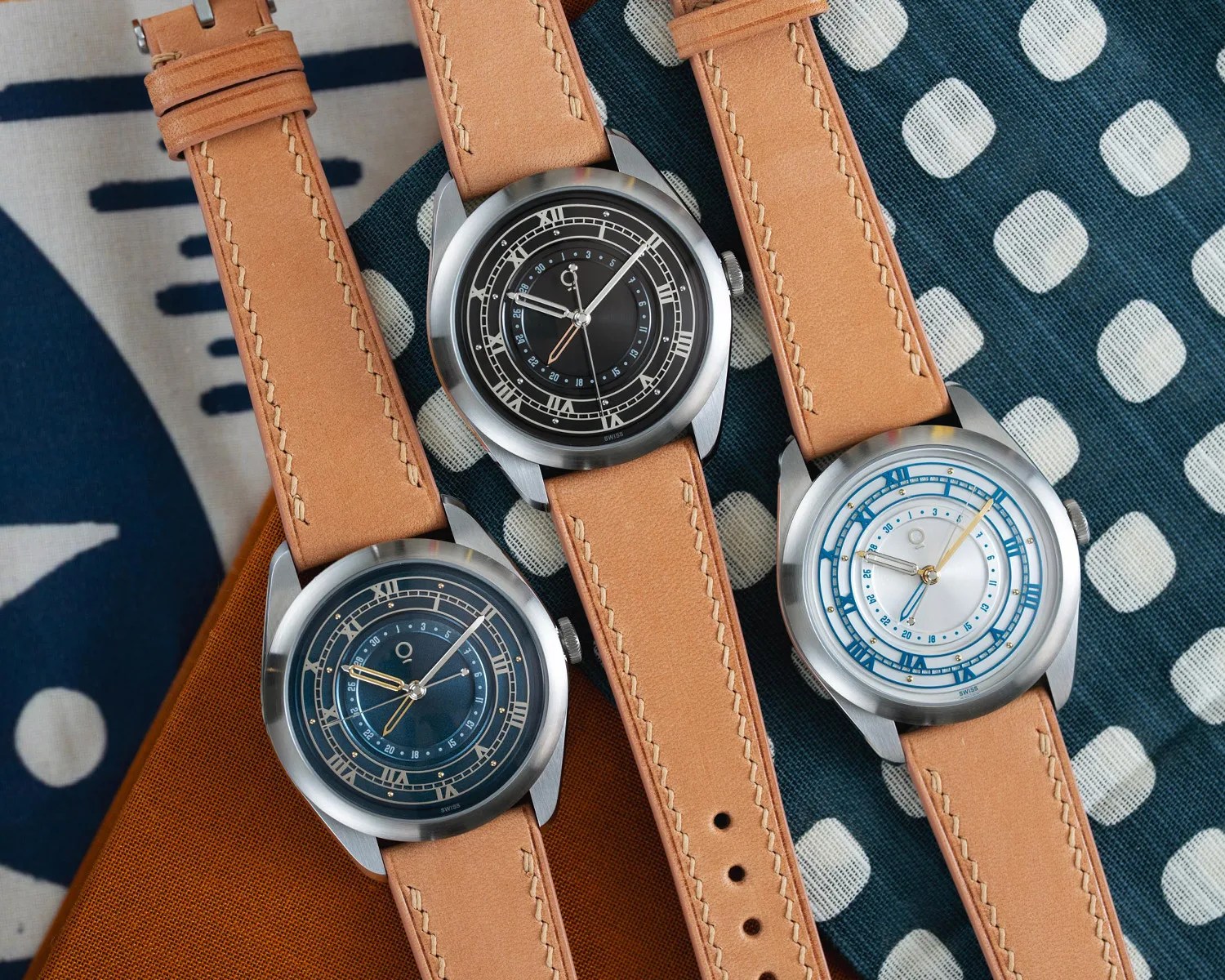 Three wristwatches with tan leather straps and round stainless steel cases, featuring black, blue, and white dials with Roman numerals.