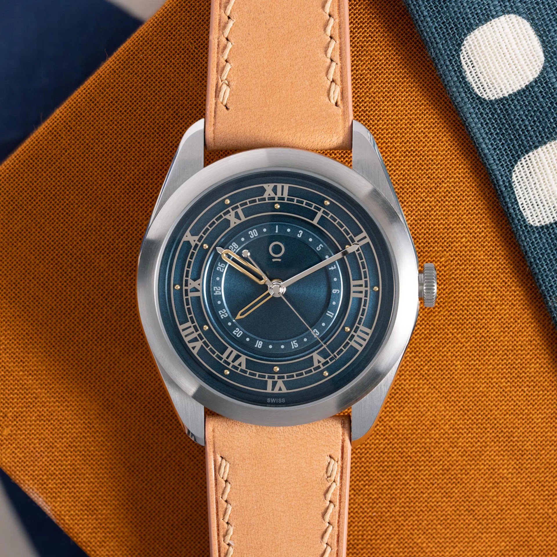 Silver wristwatch with blue dial, Roman numerals, and tan leather strap on orange and blue fabric.