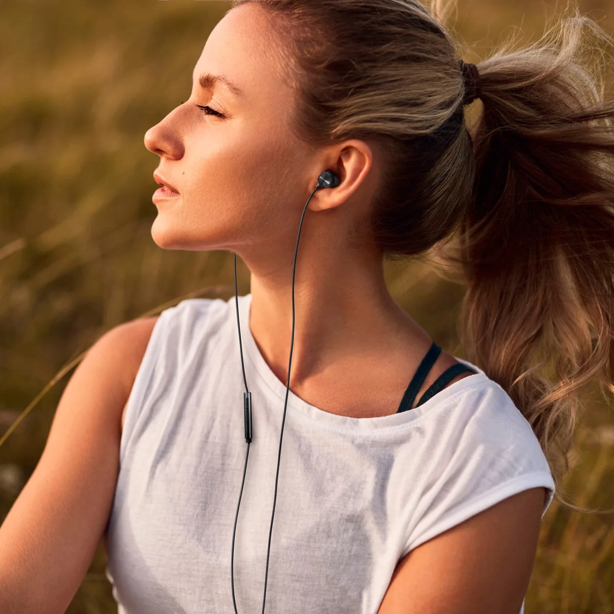 Woman with blonde ponytail wearing black wired earbuds and a white sleeveless top outdoors.