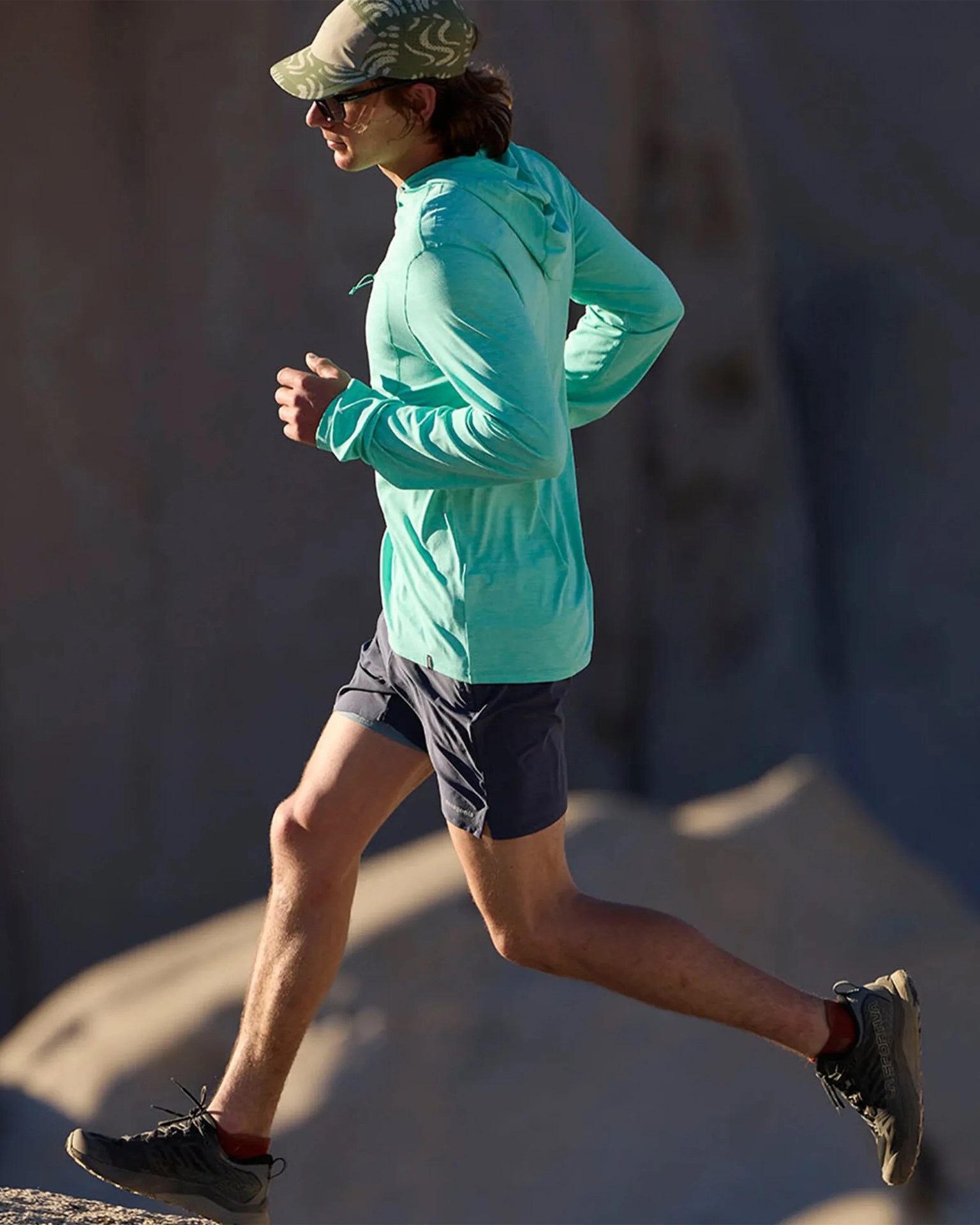 Person running outdoors wearing a light turquoise hooded long-sleeve shirt, dark shorts, and black trail running shoes.