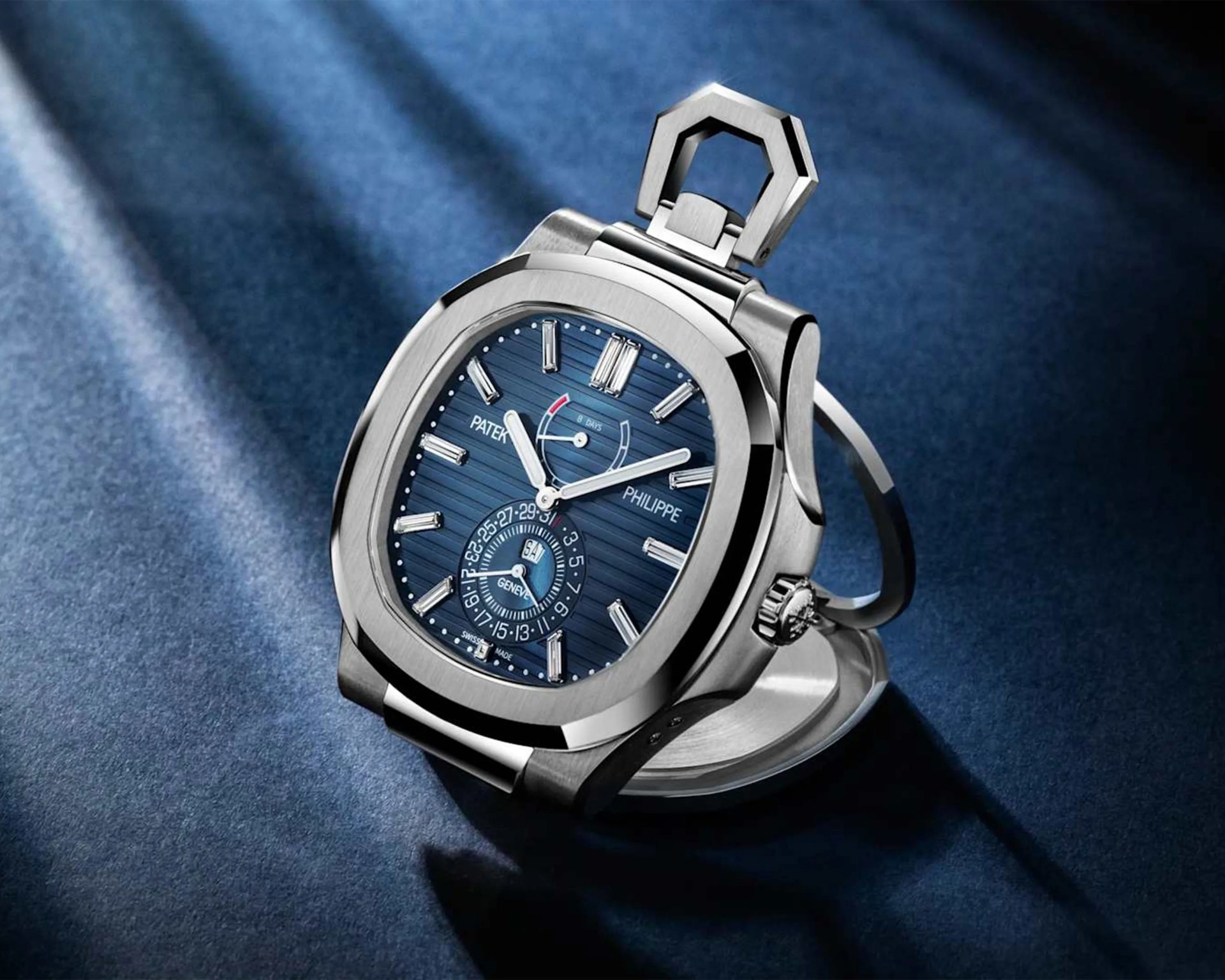 Silver Patek Philippe pocket watch with a blue dial and diamond hour markers on a blue fabric surface.