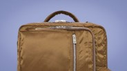 Brown nylon backpack with silver zippers and a top handle against a light purple background.