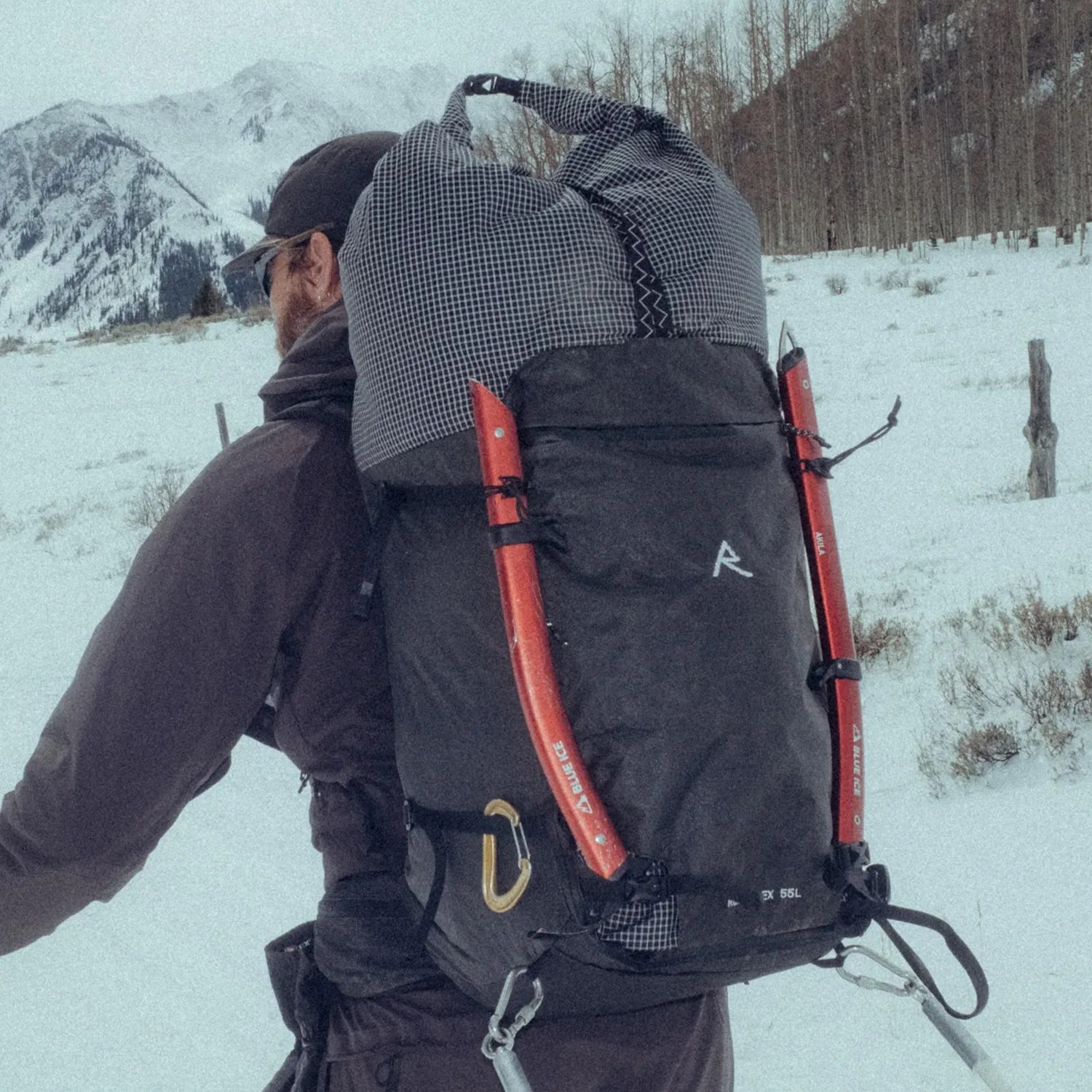 Black and gray 55L hiking backpack with red ice axes attached, worn by a person in a snowy mountain setting.