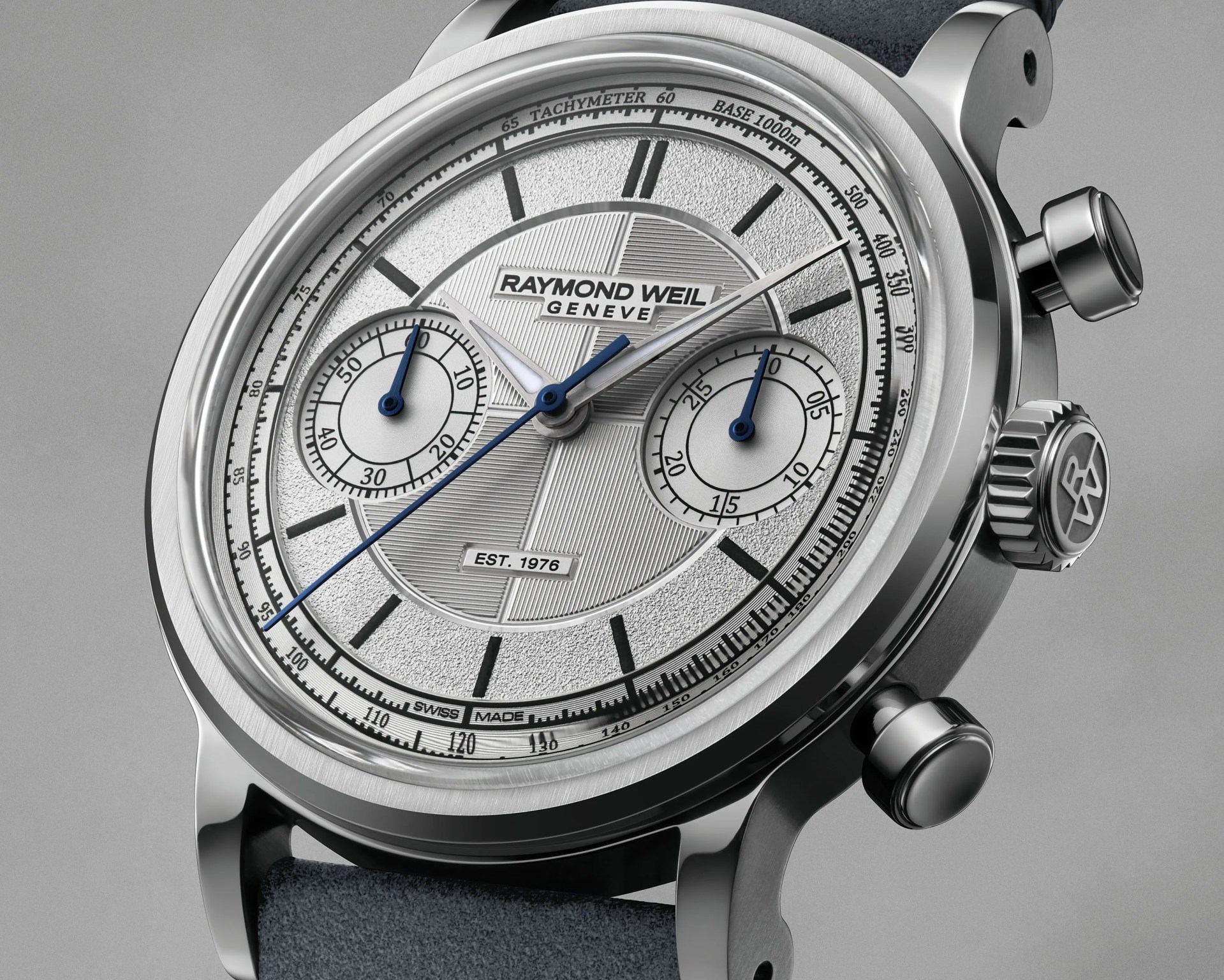 Close-up of a Raymond Weil Geneve silver chronograph watch with blue hands and tachymeter scale on a gray background.