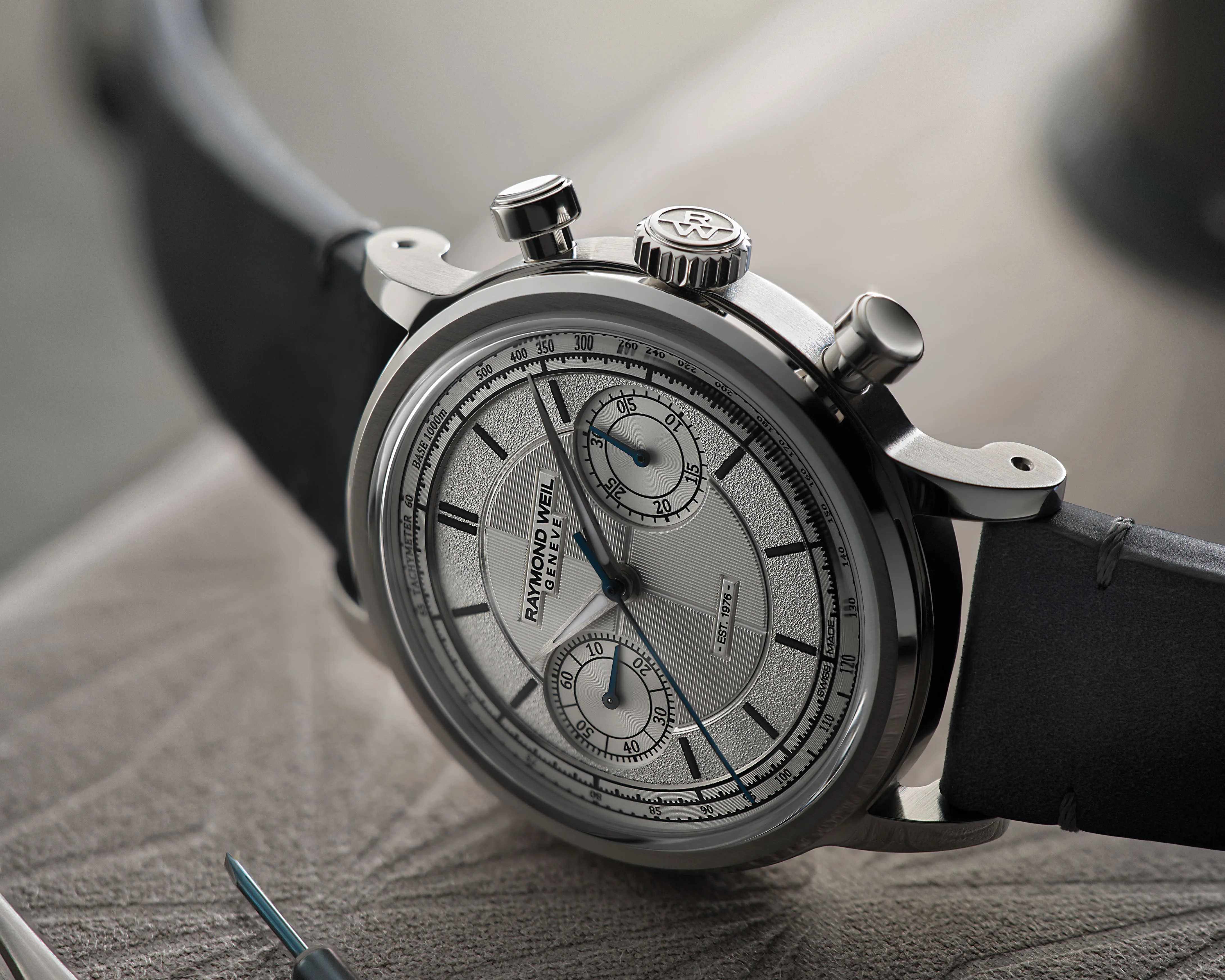 Silver Raymond Weil chronograph watch with black leather strap on textured surface.