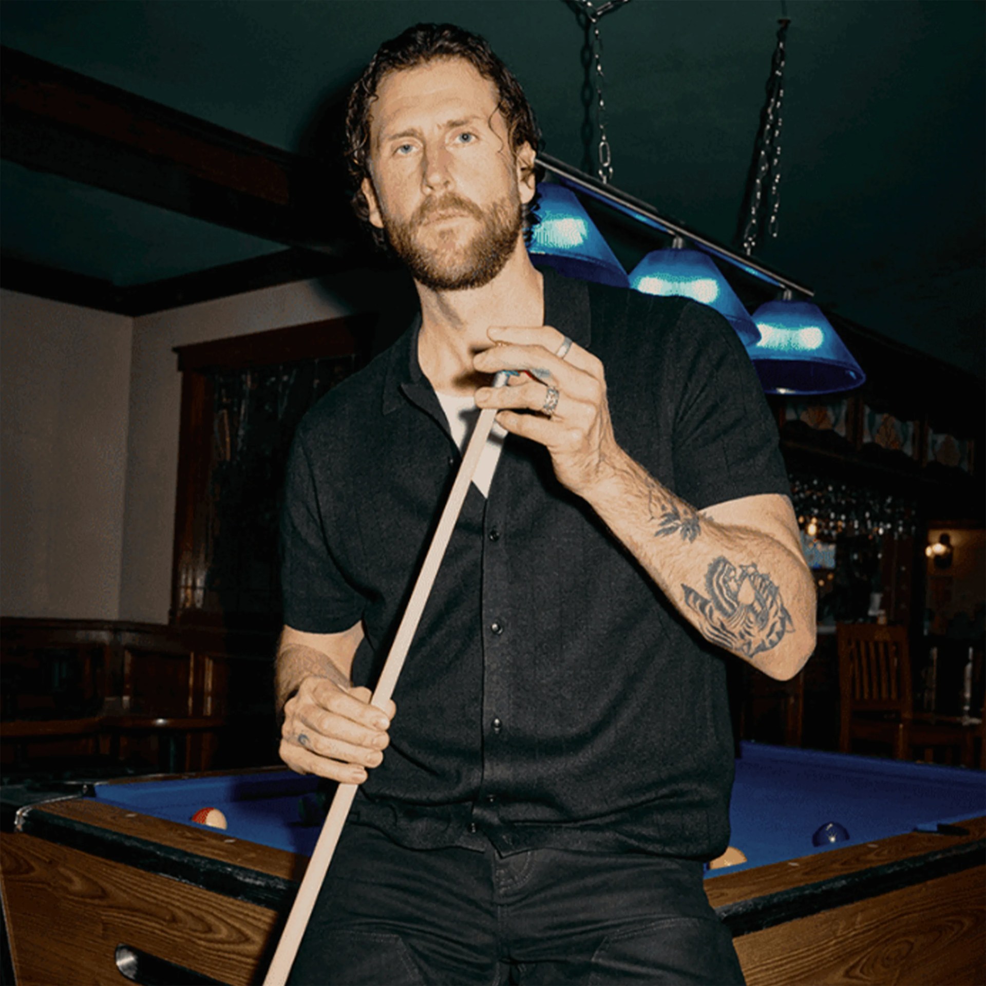 Man with tattoos wearing a black short-sleeve button-up shirt holding a pool cue near a pool table.