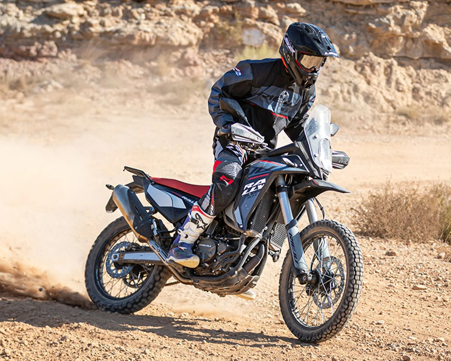 Black and gray off-road motorcycle with red seat ridden on a dirt trail by a person in black gear and helmet.