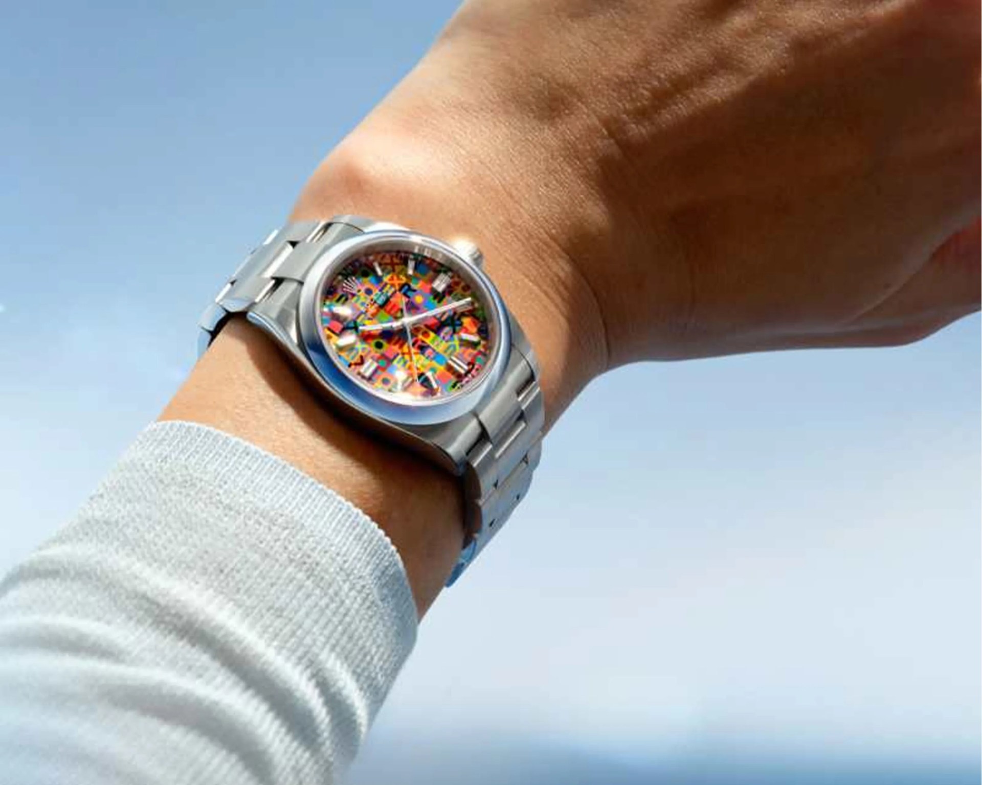Silver metal wristwatch with a colorful abstract patterned dial worn on a wrist against a blue sky background.