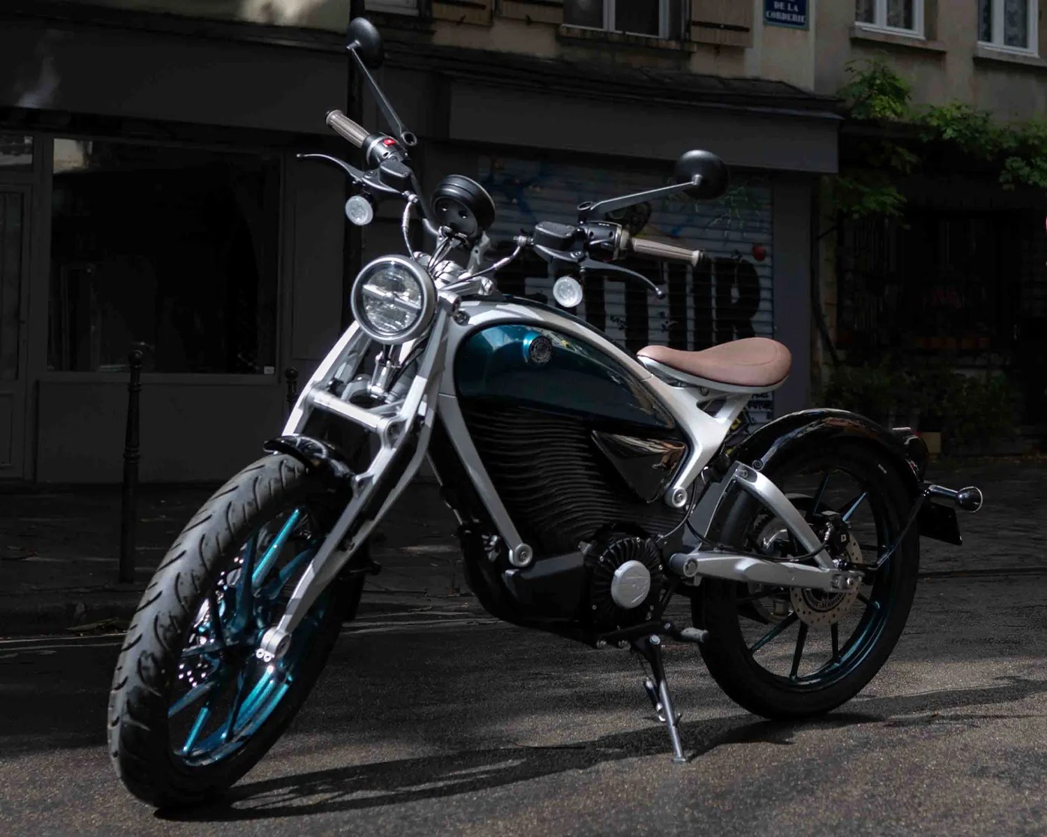 Electric motorcycle with teal wheels, silver frame, and tan seat parked on a street.
