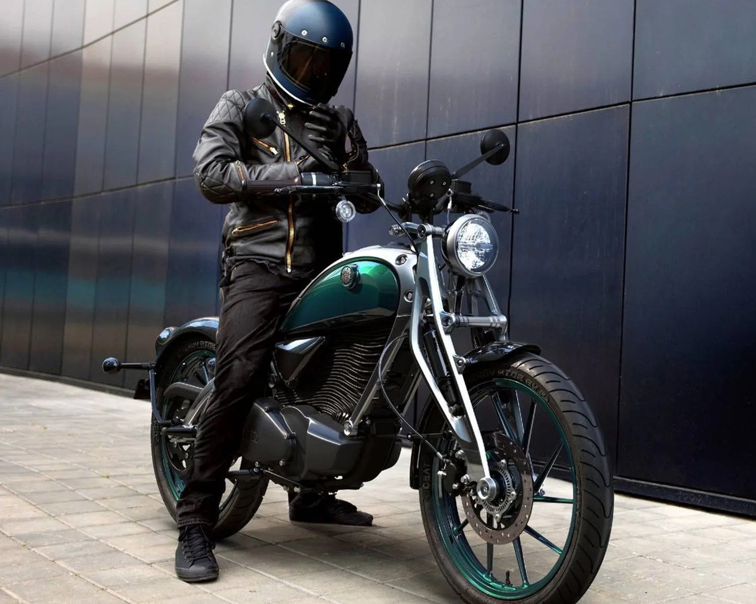 Person in black leather jacket and blue helmet sitting on a black and green motorcycle with a round headlight.