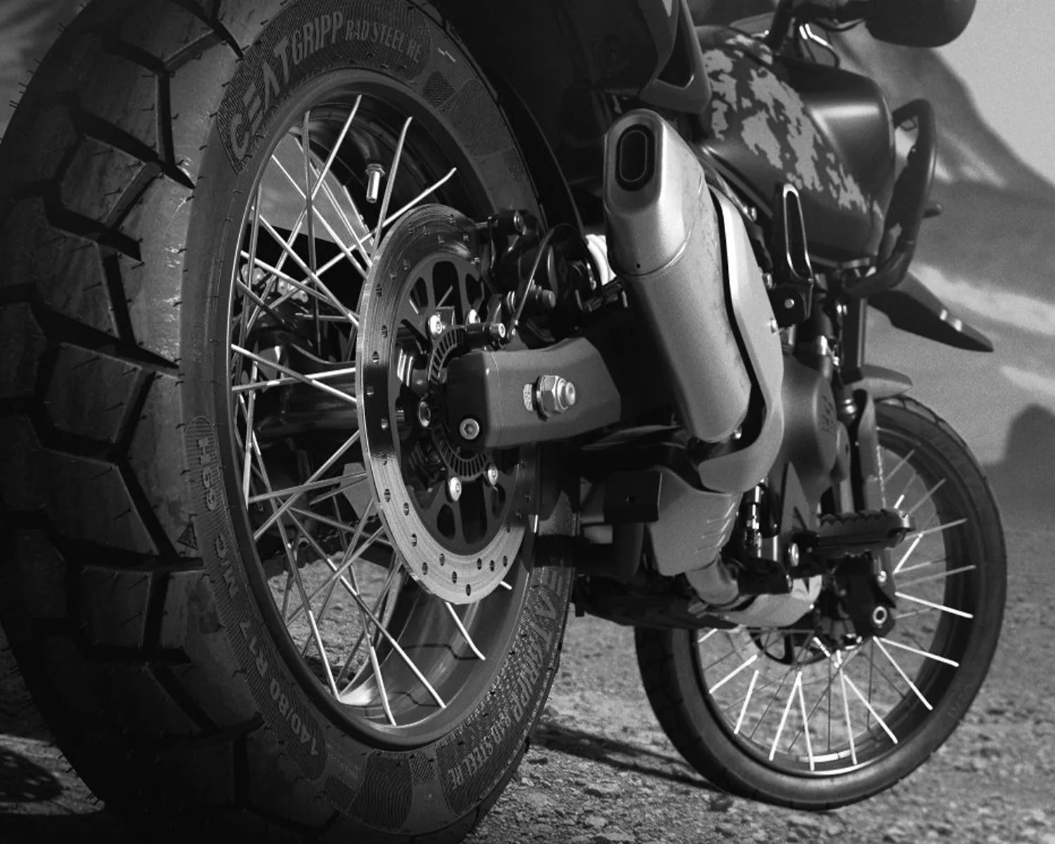 Close-up of a motorcycle's rear and front wheels with spoked rims and knobby tires on a gravel surface.
