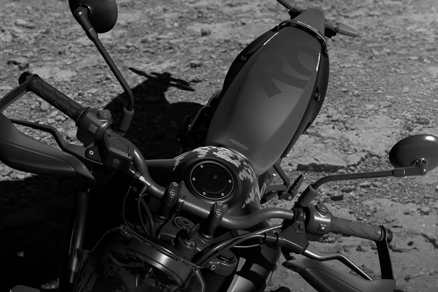Top view of a matte black Royal Enfield motorcycle fuel tank and handlebars on a rough gravel surface.