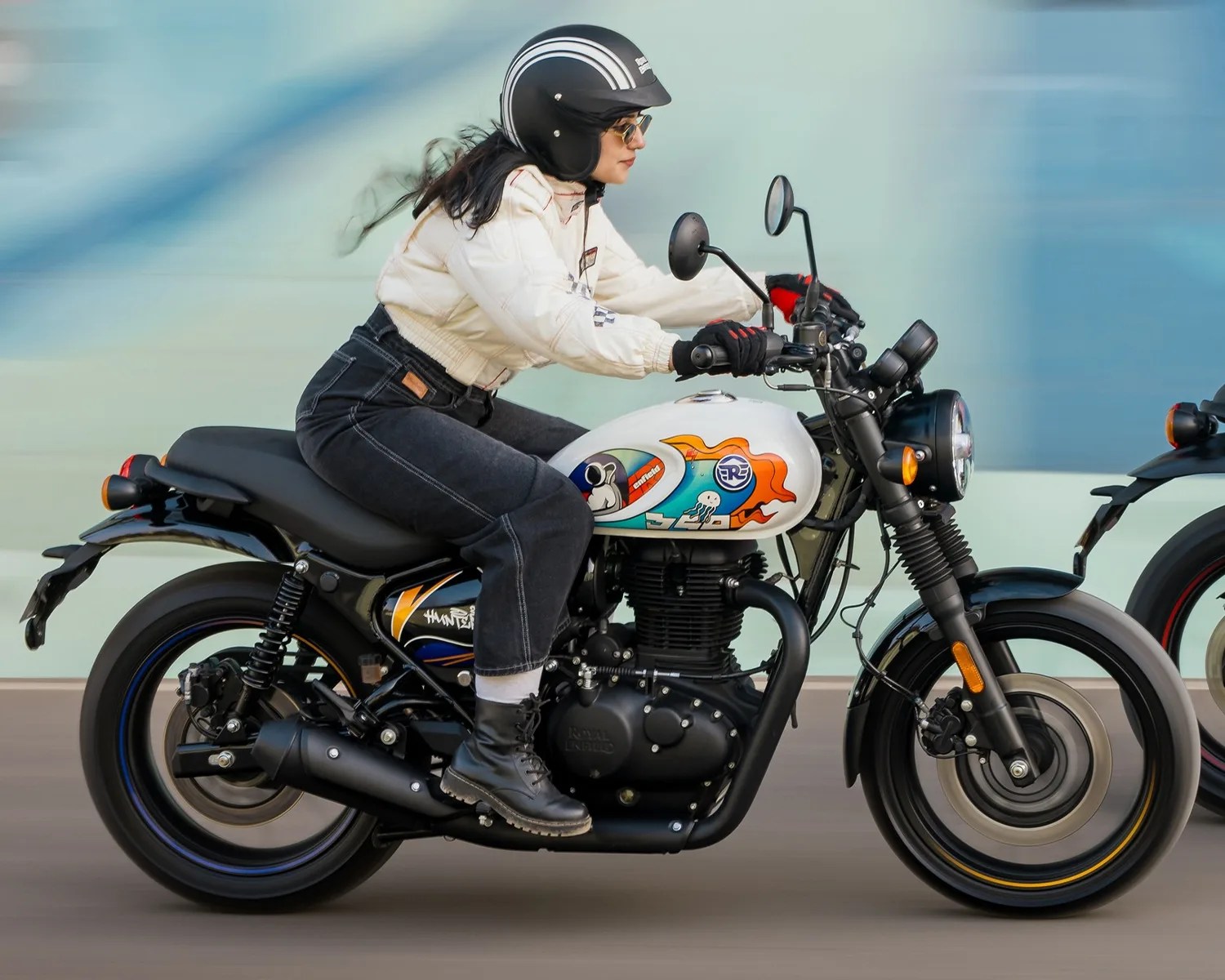 Person wearing a black helmet and white jacket riding a black and white Royal Enfield motorcycle with colorful tank graphics.