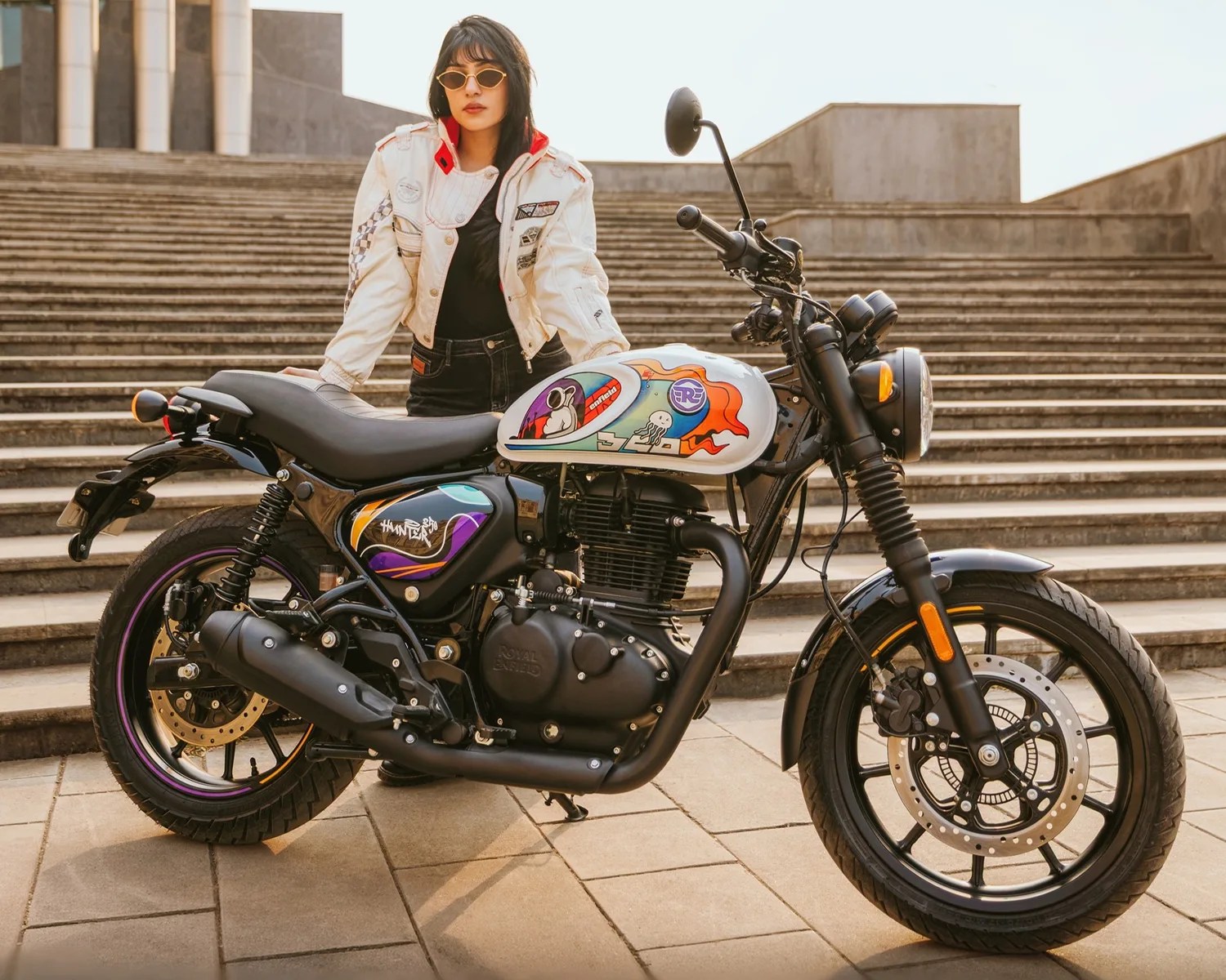 Black and white Royal Enfield Hunter 350 motorcycle with colorful tank art parked on pavement in front of stone steps.