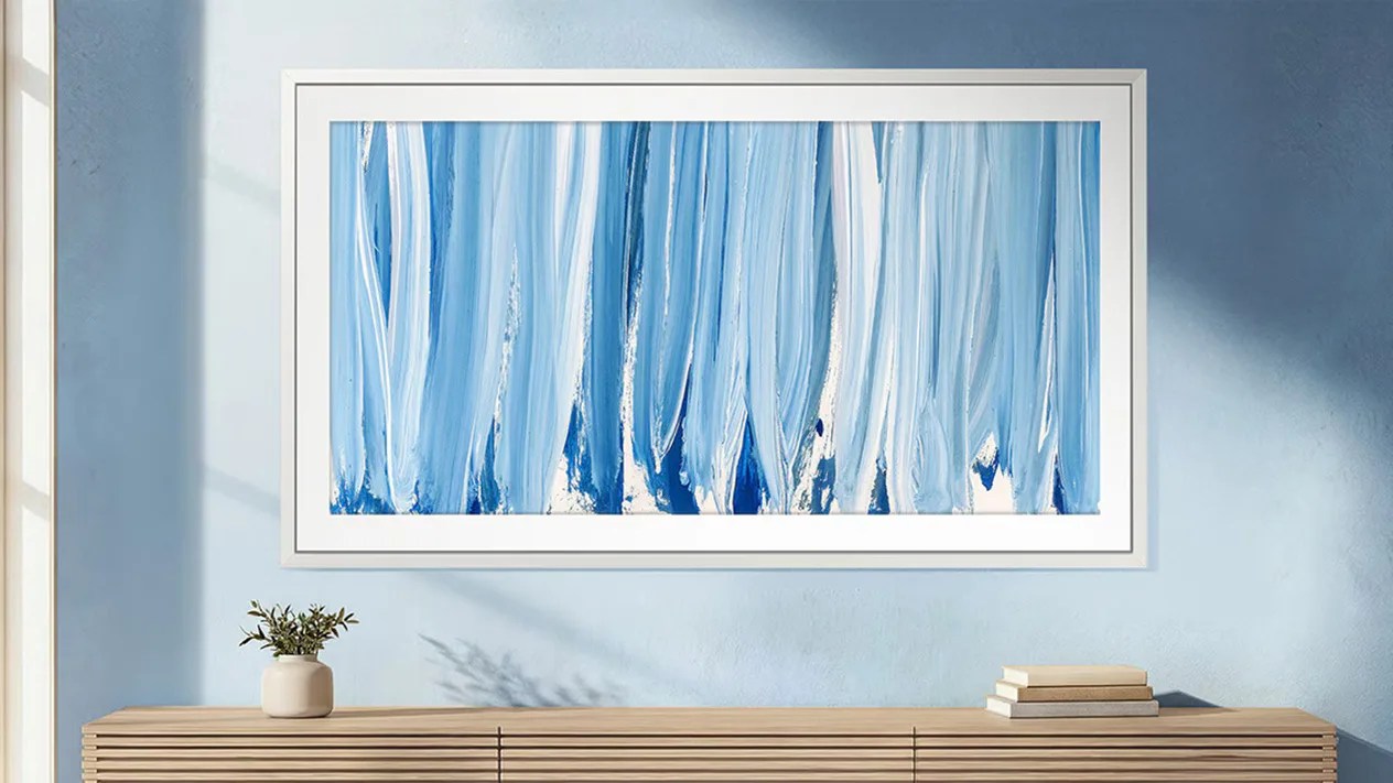 Abstract blue and white vertical brushstroke painting in a white frame on a light blue wall above a wooden sideboard with a small plant and stacked books.