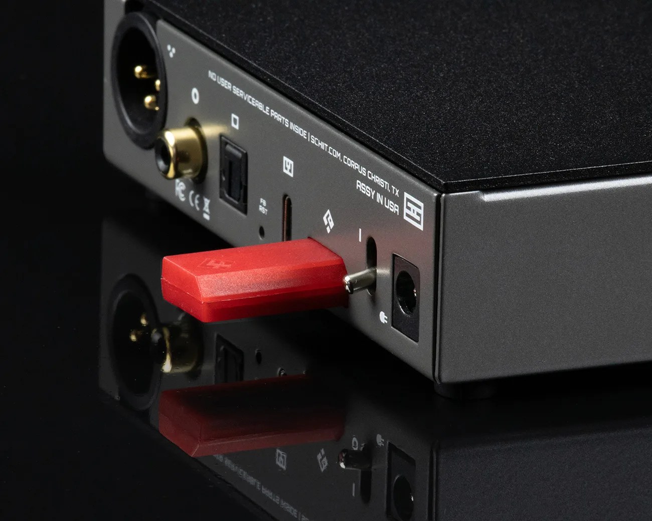 Black audio device with gold connectors and a red USB stick plugged into the back panel on a reflective surface.