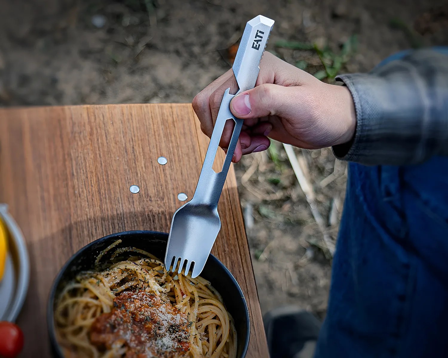 Hand holding a silver metal spork with "EATi" engraved, above a bowl of spaghetti on a wooden table.