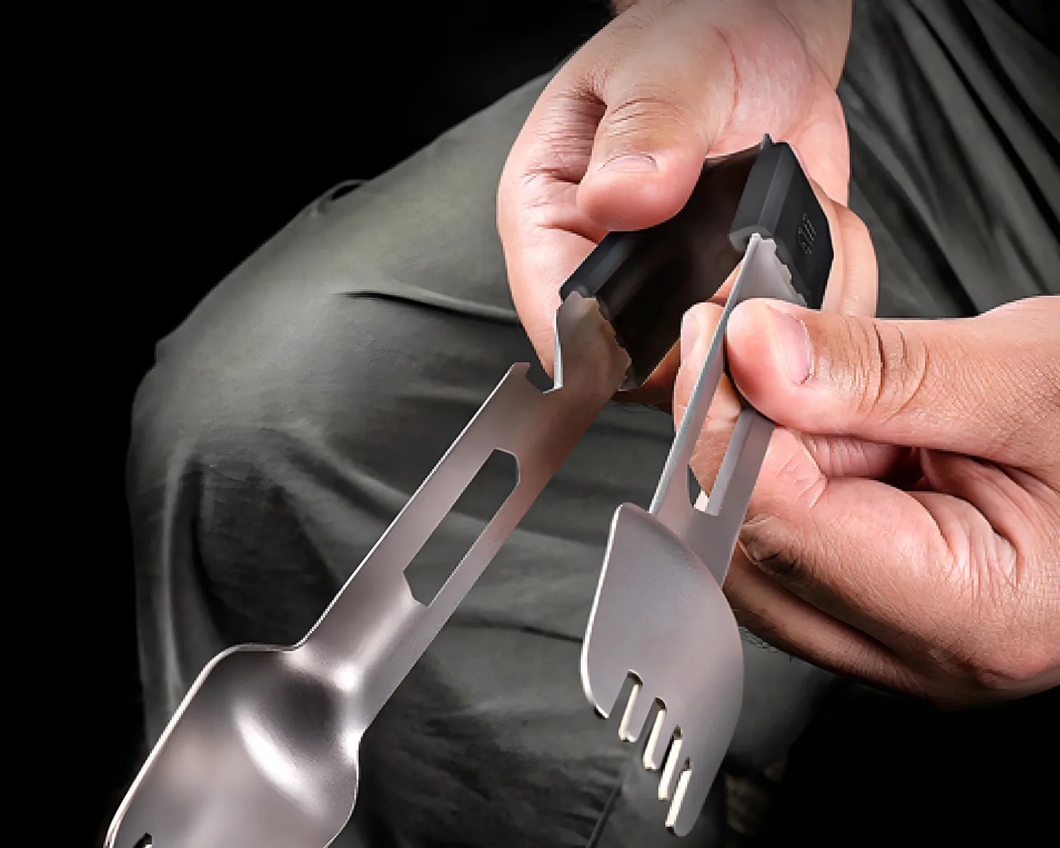 Hands holding a compact, foldable stainless steel spork with a black plastic hinge.