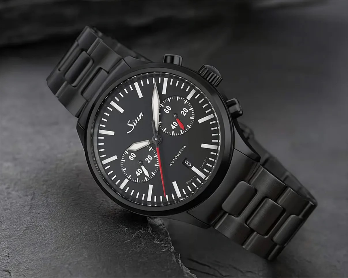 Black Sinn wristwatch with metal link band, black dial, white markers, two subdials, and red second hand on a dark surface.