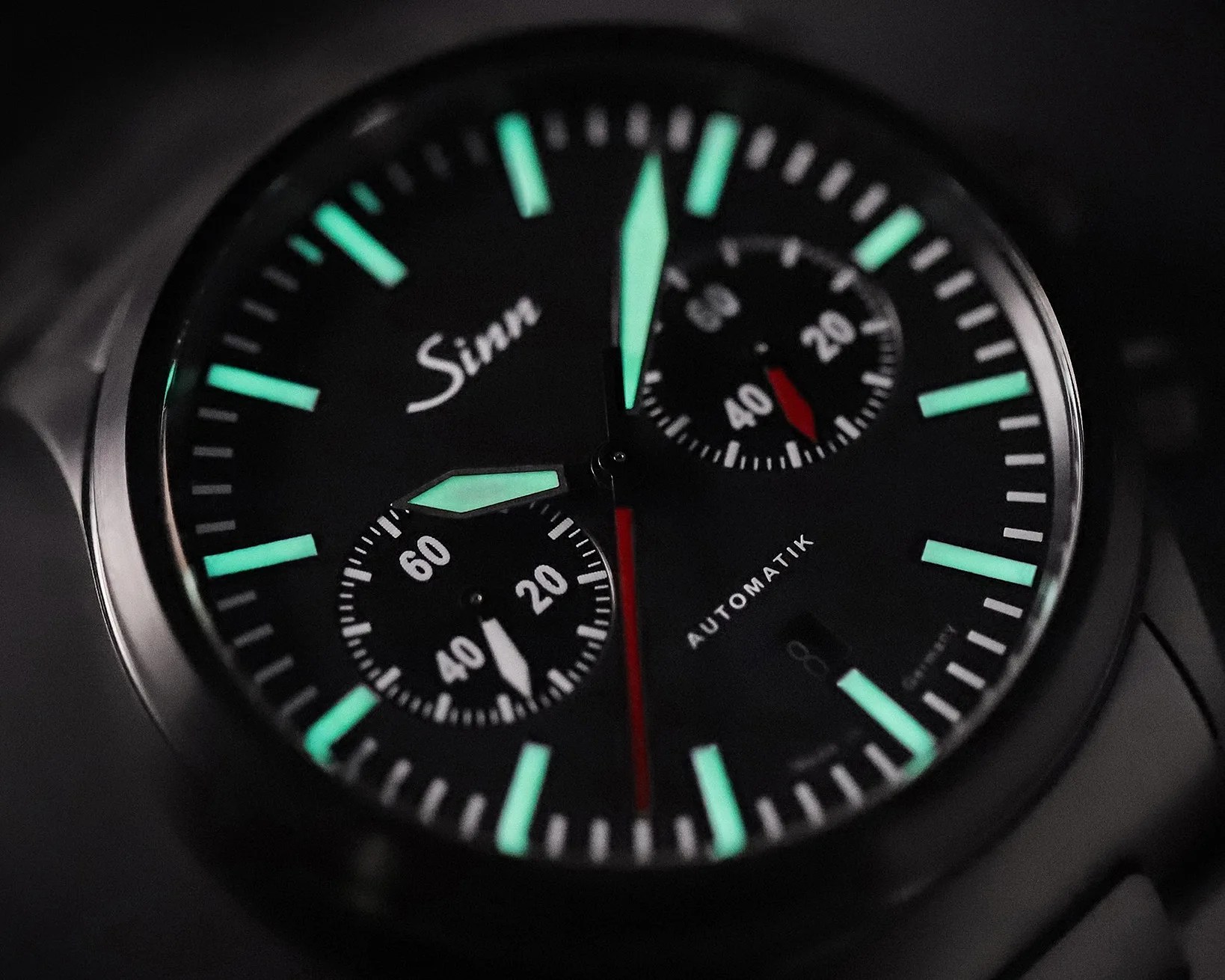 Close-up of a black Sinn wristwatch dial with luminous green hour markers and hands, two subdials, and a red second hand.