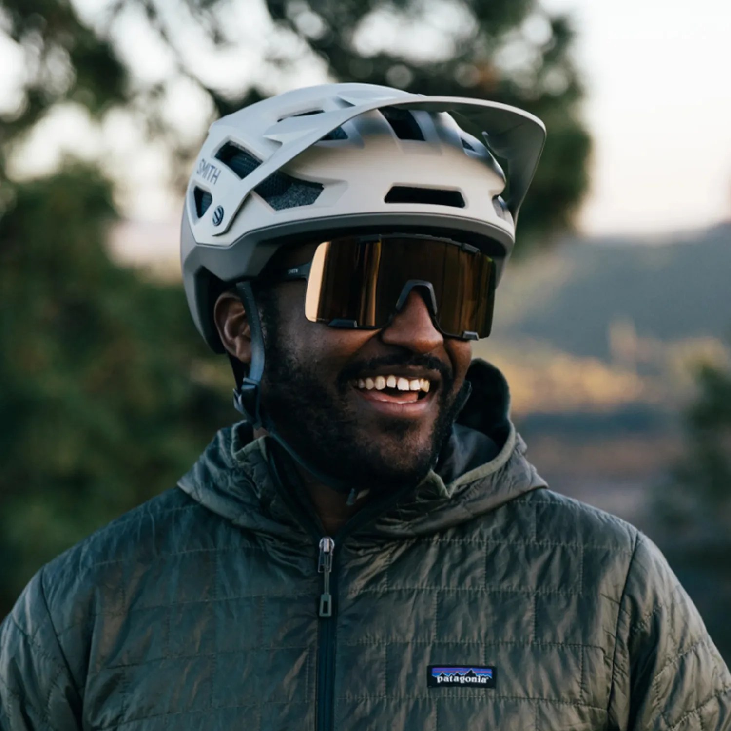 Man wearing a white Smith bike helmet, brown reflective sunglasses, and a green Patagonia jacket outdoors.